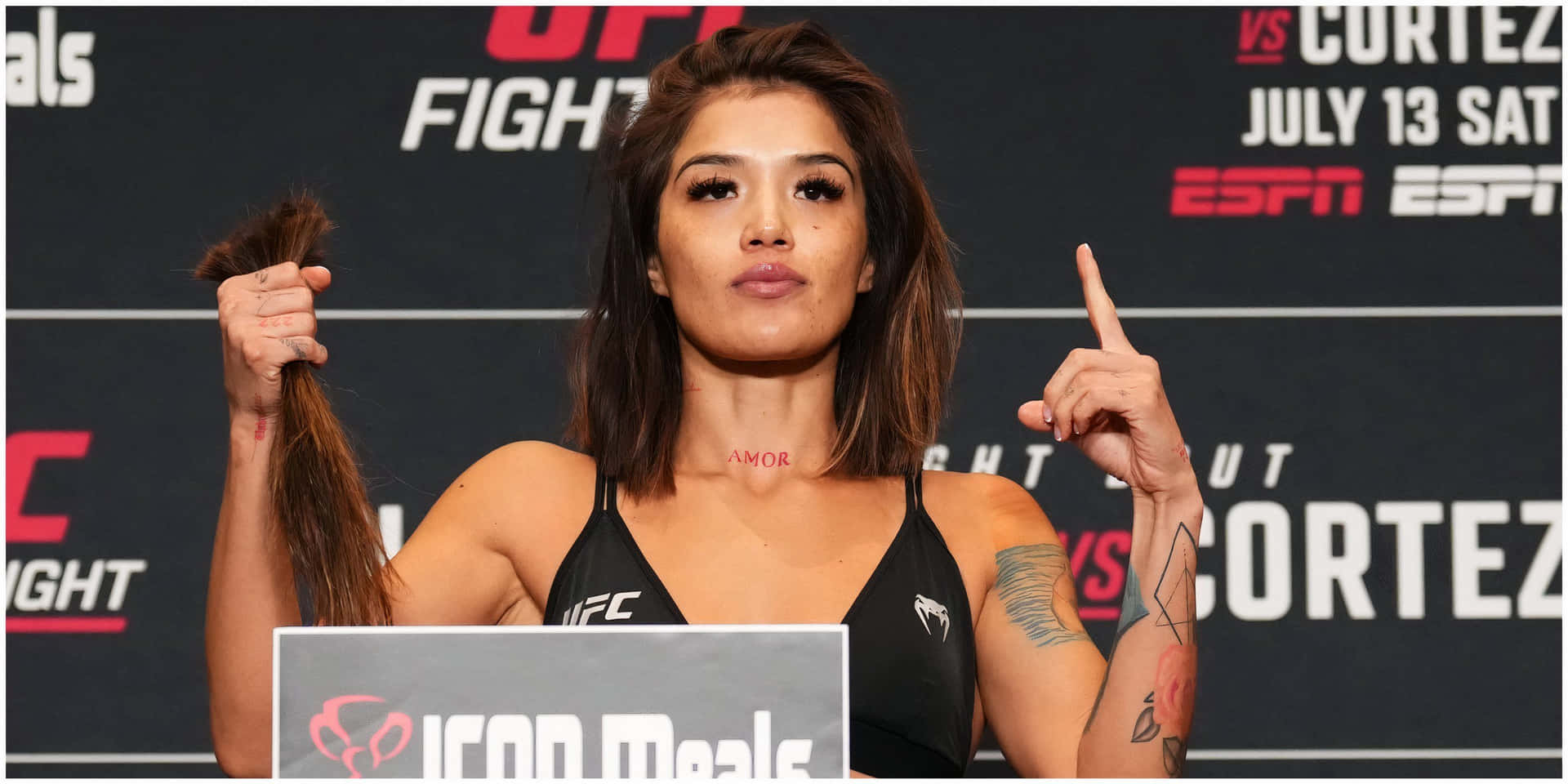  壁紙 — 一位女性UFC拳手自信地站在稱重台上，握著馬尾辮並向上指，手臂上的紋身清晰可見。