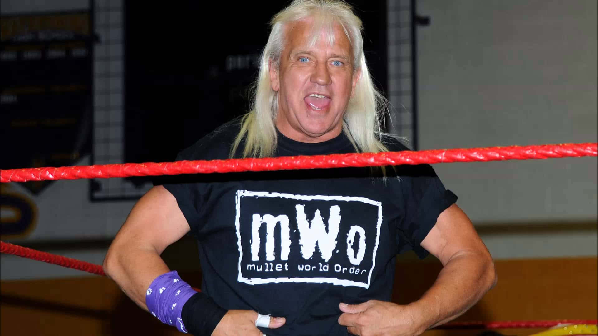 前 NWA 重量級冠軍 Ricky Morton 壁紙 — 一位肌肉發達的摔角手，留著金色長髮，戴著紫色腕帶，在紅色邊框的摔角場內自信地擺姿勢。