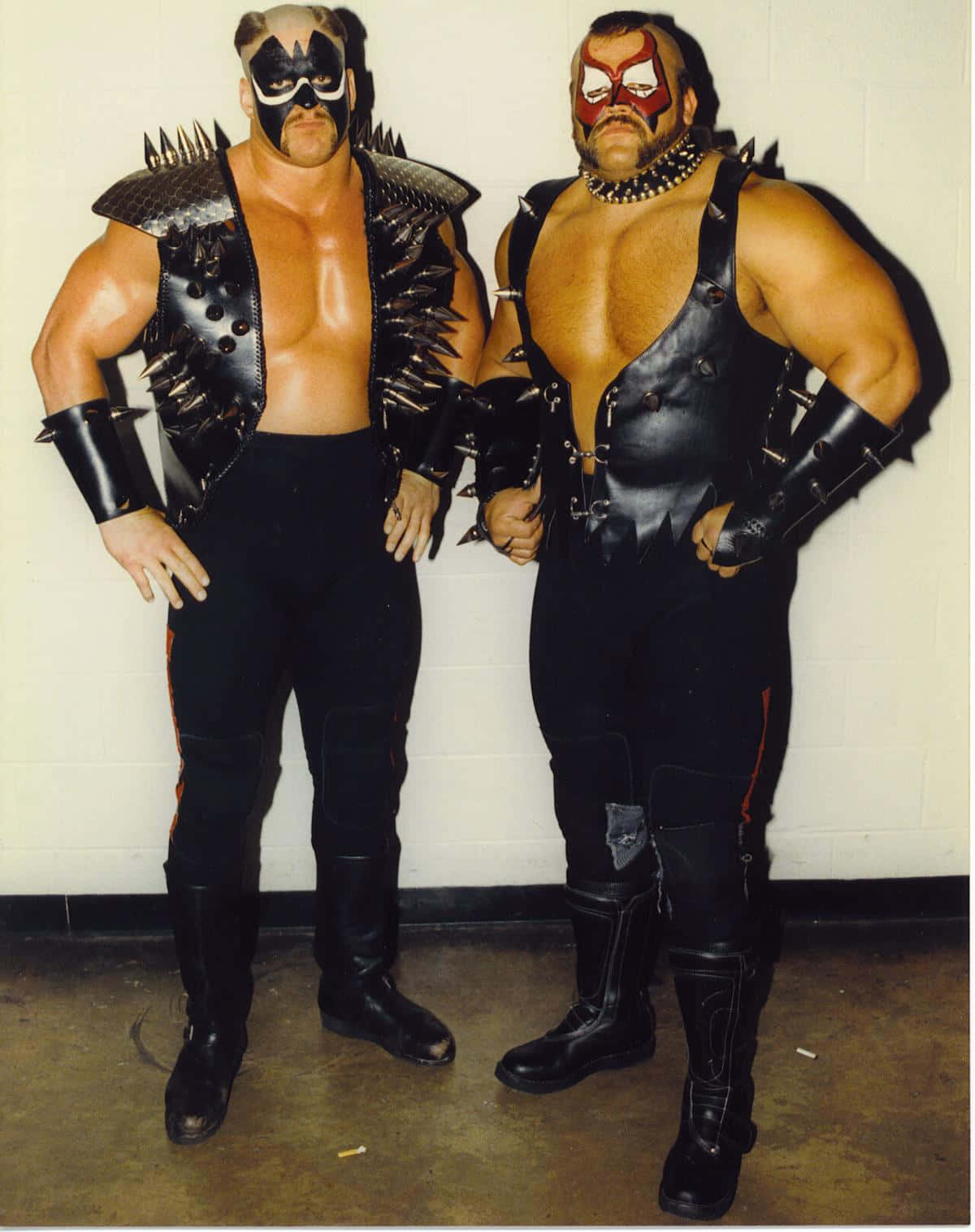 NWA冠軍Road Warrior Hawk與Road Warrior Animal激戰時刻 壁紙 — 兩名肌肉發達的摔角手穿著黑色釘皮裝備和臉部彩繪，自信地靠著素牆擺姿勢。