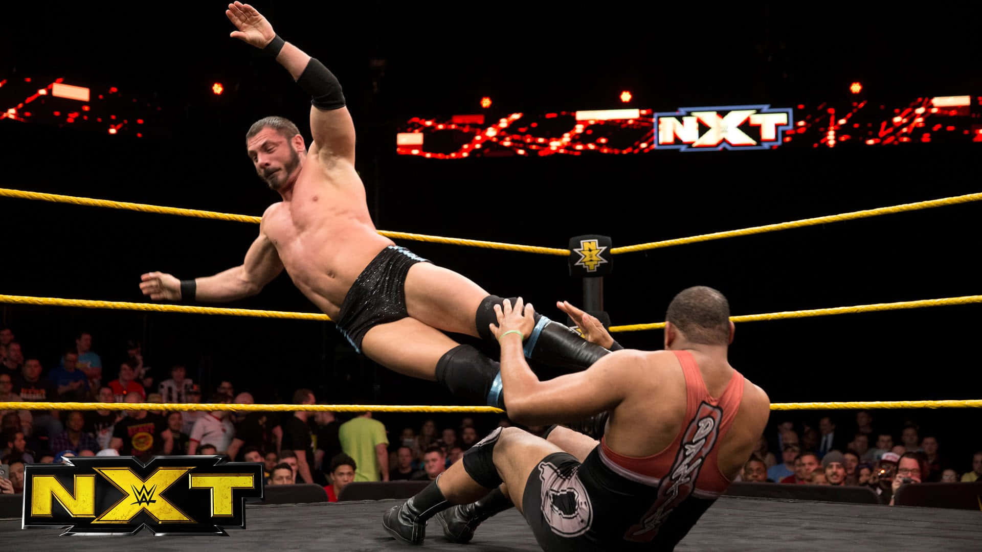 奧斯汀·艾瑞斯 奪取 NXT 比賽主導權 壁紙 — 一名摔角手在燈光明亮的NXT擂台上躍向對手，背景是熱情歡呼的粉絲。