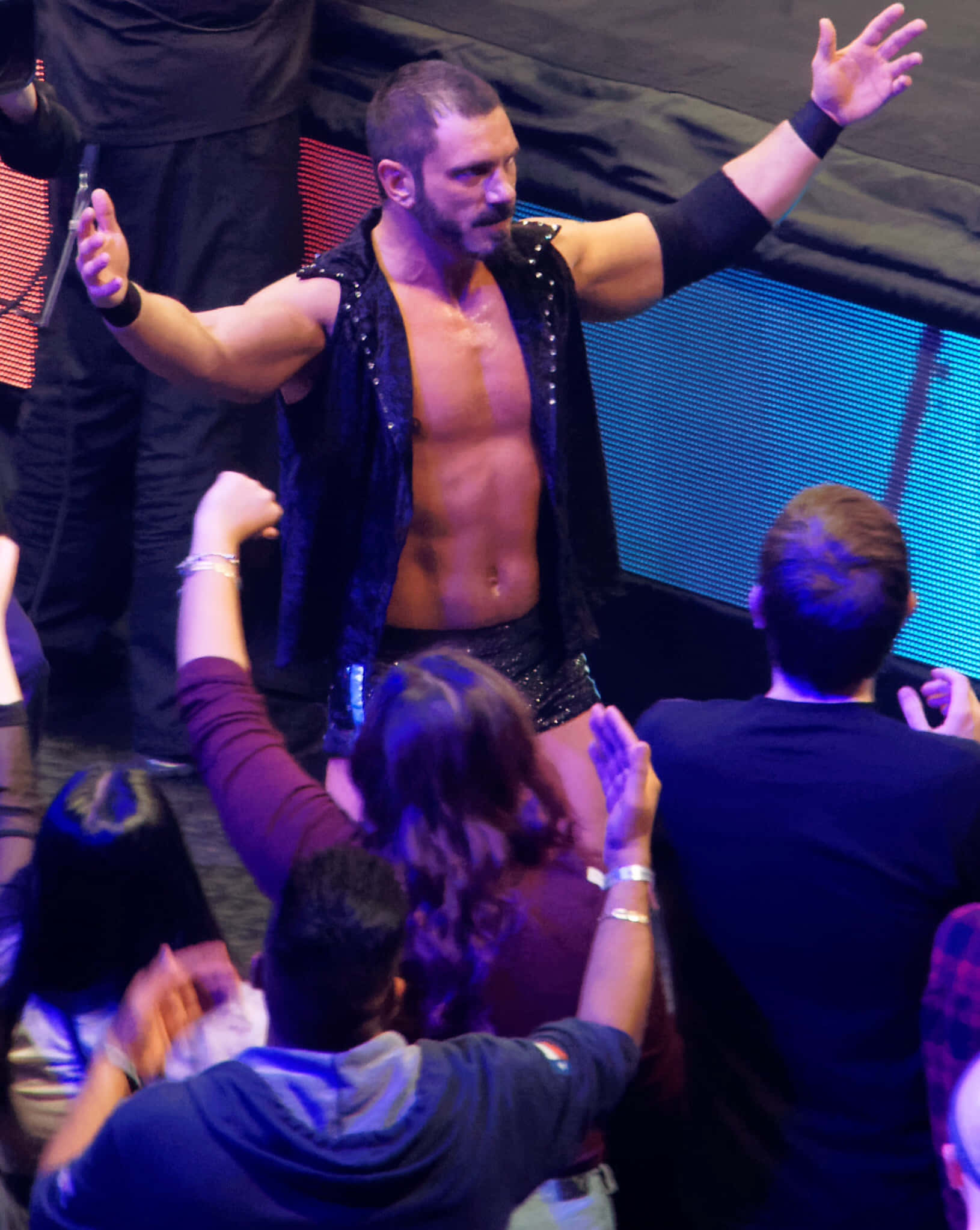 NXT Takeover 奧斯汀·艾瑞斯 壁紙 — 一位肌肉發達的摔角手穿著黑色閃片背心，在藍色舞台燈光下高舉雙臂，粉絲們伸手觸碰他，場面勝利輝煌。