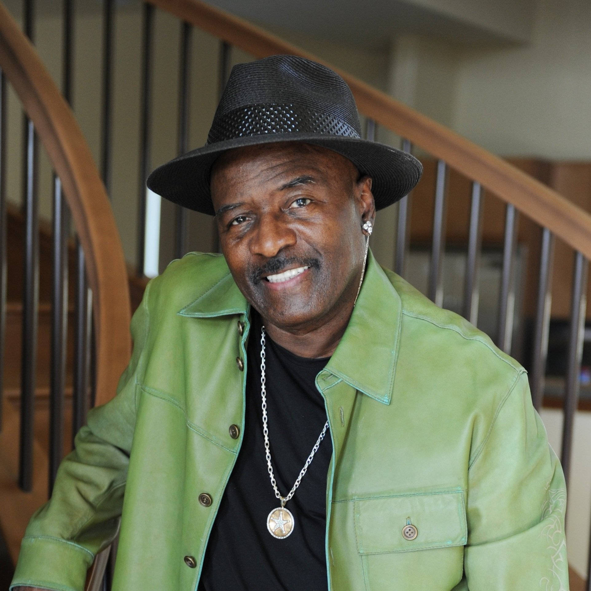 O'Jays Walter Williams 演唱會現場 壁紙 — 一位微笑的男子身穿綠色皮夾克和黑色軟呢帽，在室內木樓梯附近擺姿勢。