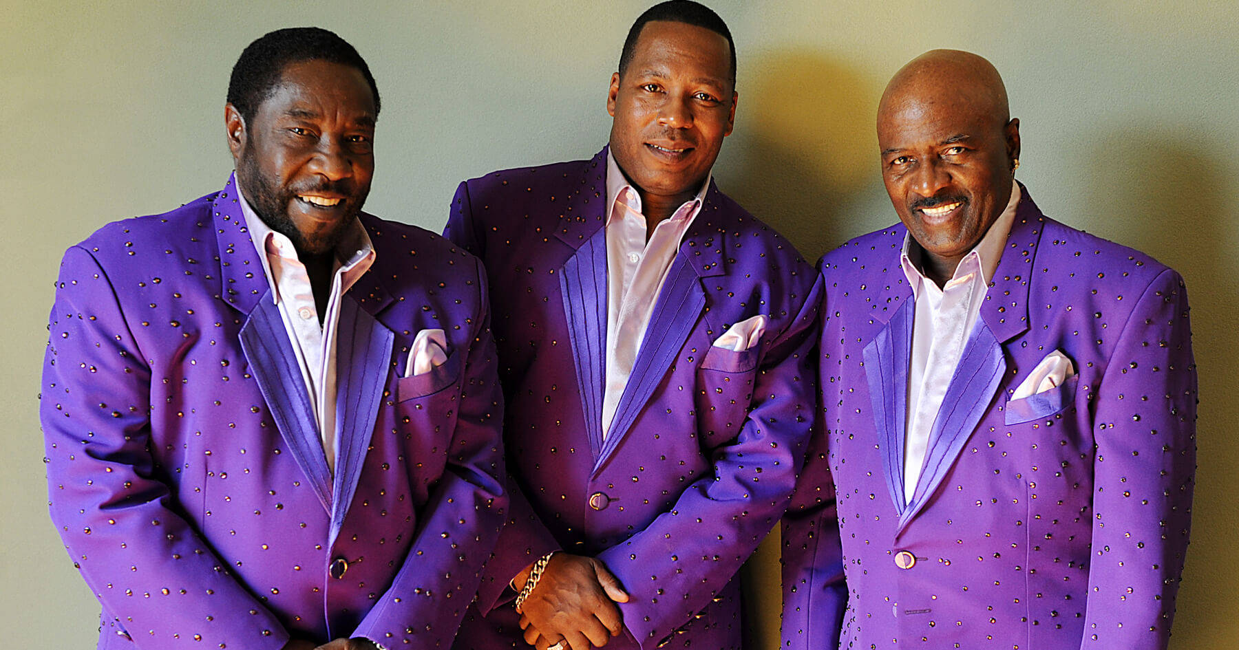 O'Jays 傳奇音樂演出於亞利桑那 PBS 壁紙 — 三個微笑的男人穿著相配的紫色亮片西裝，自信地靠著素牆擺姿勢。