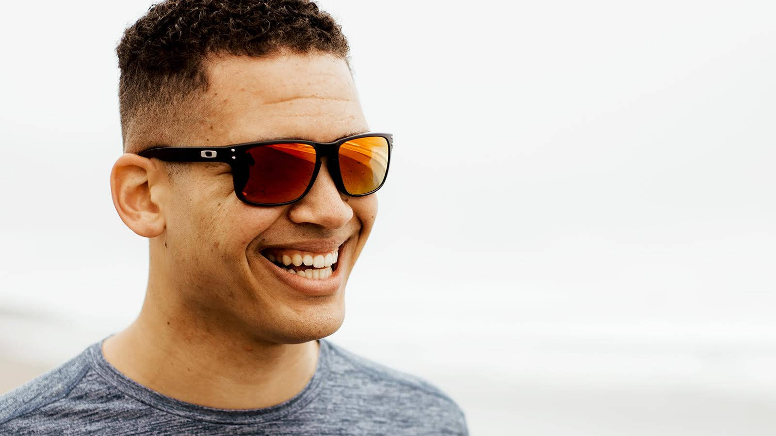 Oakley Holbrook 偏光太陽眼鏡時尚款 壁紙 — 一個微笑的捲髮男子戴著黑色太陽眼鏡，橙色鏡片，背景是明亮的白色。