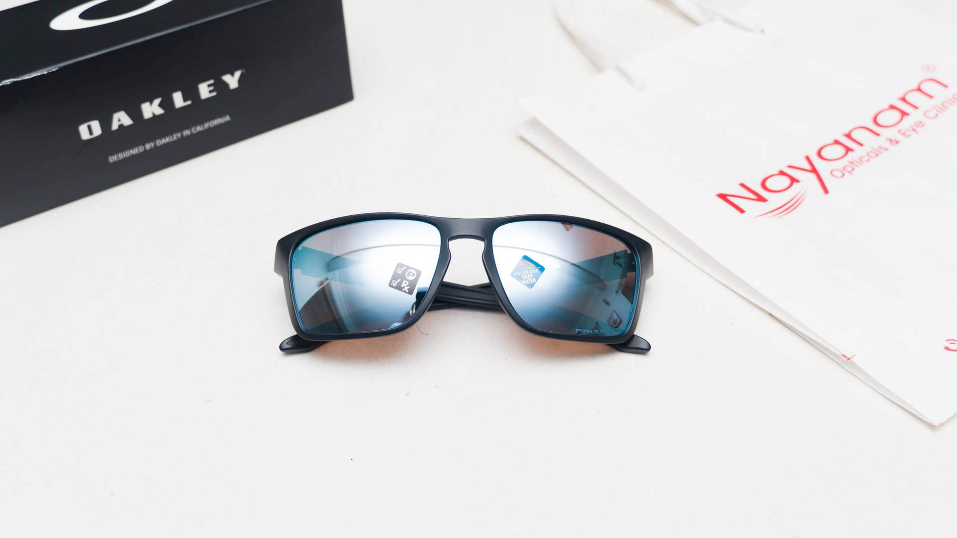 Oakley Holbrook運動冒險太陽眼鏡 壁紙 — 黑色Oakley太陽眼鏡配藍色鏡面鏡片，放在白色表面上，旁邊有盒子和Nayaram手袋。