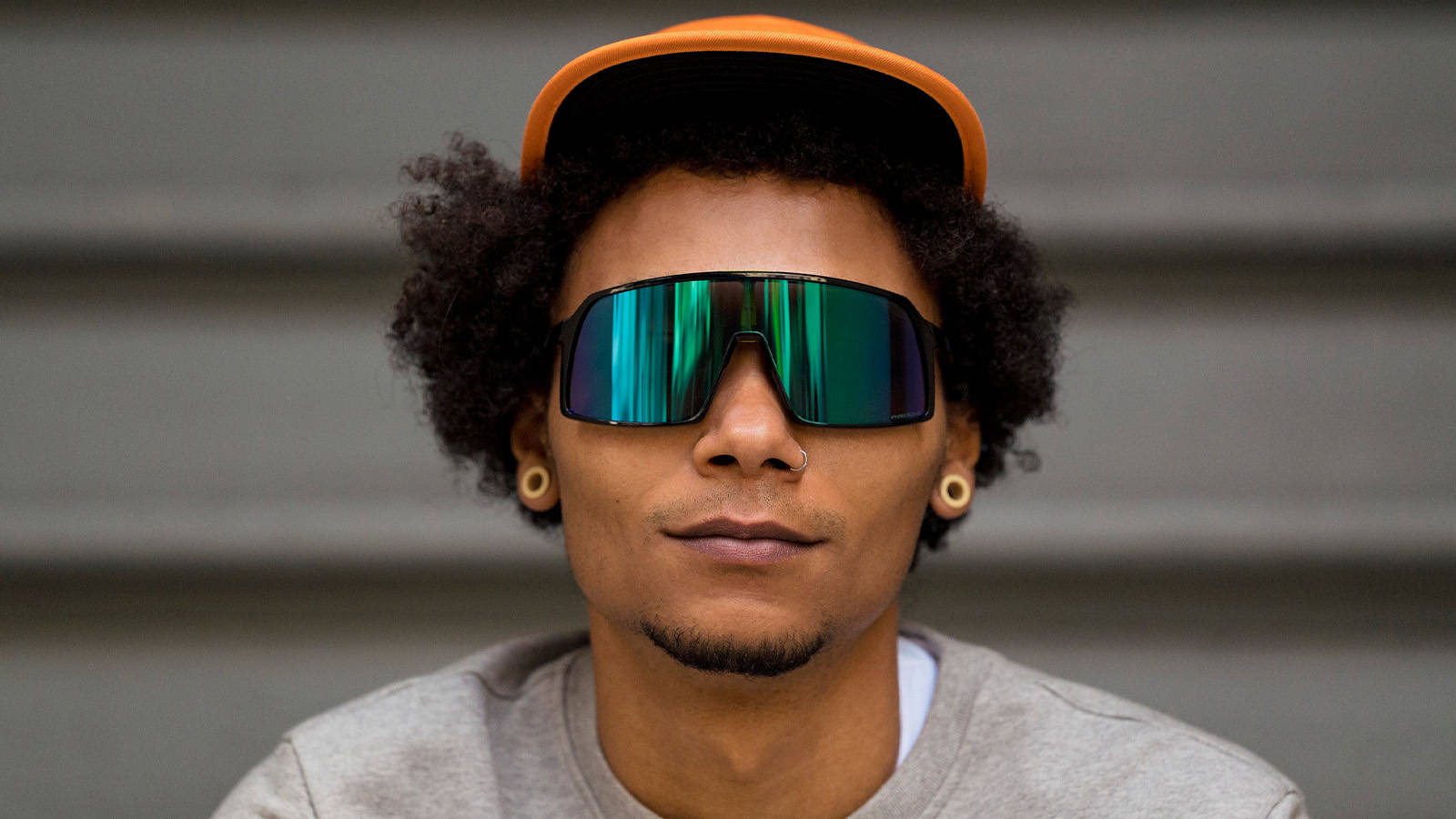 Oakley Sutro 運動性能太陽眼鏡 壁紙 — 一個時尚嘅人，有曲髮，戴住搶眼嘅橙色帽同反光綠色太陽眼鏡，背景係灰色嘅瓦楞紙。