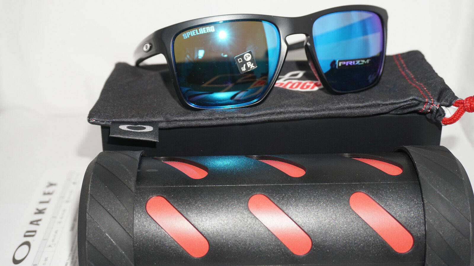 Oakley 男士冒險運動太陽眼鏡 壁紙 — 黑色Oakley太陽眼鏡，藍色鏡面鏡片，放在品牌眼鏡盒和紅色點綴的硬殼眼鏡盒上。
