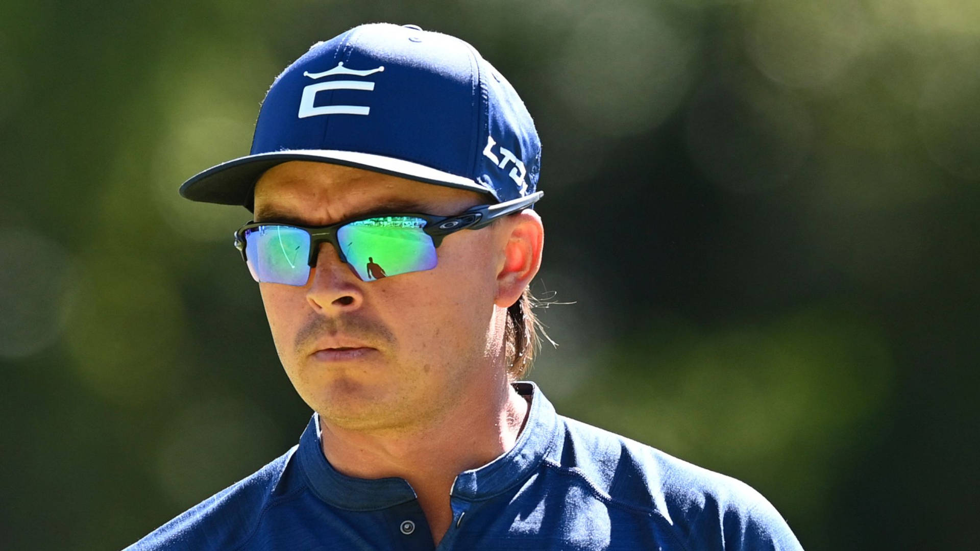 Oakley 男士運動太陽眼鏡 壁紙 — 一位專注的高爾夫球手戴著藍色帽子和太陽眼鏡，在明亮的陽光下站在模糊的綠色背景前。
