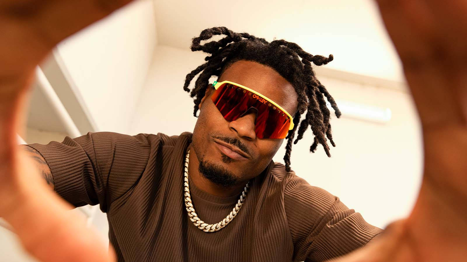 Oakley 高品質太陽眼鏡展示 壁紙 — 一個留著辮子頭、戴紅色太陽眼鏡的男人，在明亮的室內環境中，自信地透過圓形框架擺姿勢。
