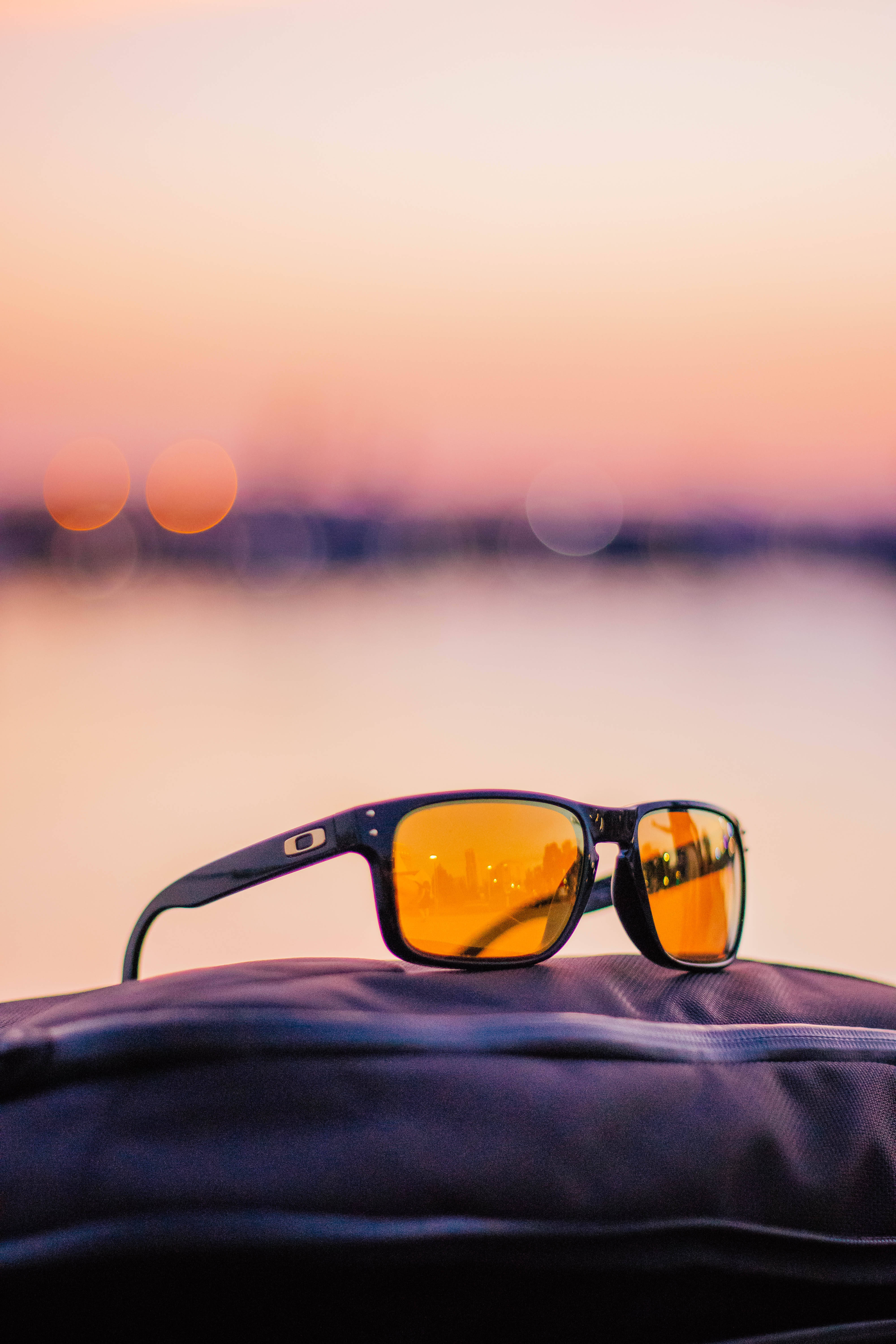 Oakley太陽眼鏡日落運動冒險 壁紙 — 琥珀色鏡片的太陽眼鏡放在深色手袋上，背景是水上柔和粉紅色的日落和散景燈光。
