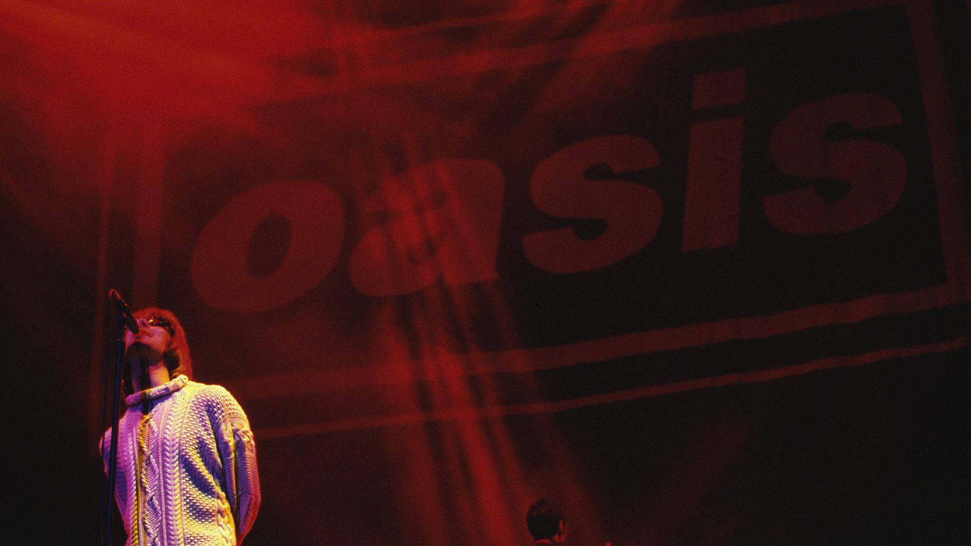 Oasis 主唱演唱 壁紙 — 一位身穿圖案襯衫的音樂家在紅色舞台燈光下對著麥克風唱歌，身後有一個巨大的「oasis」橫幅。