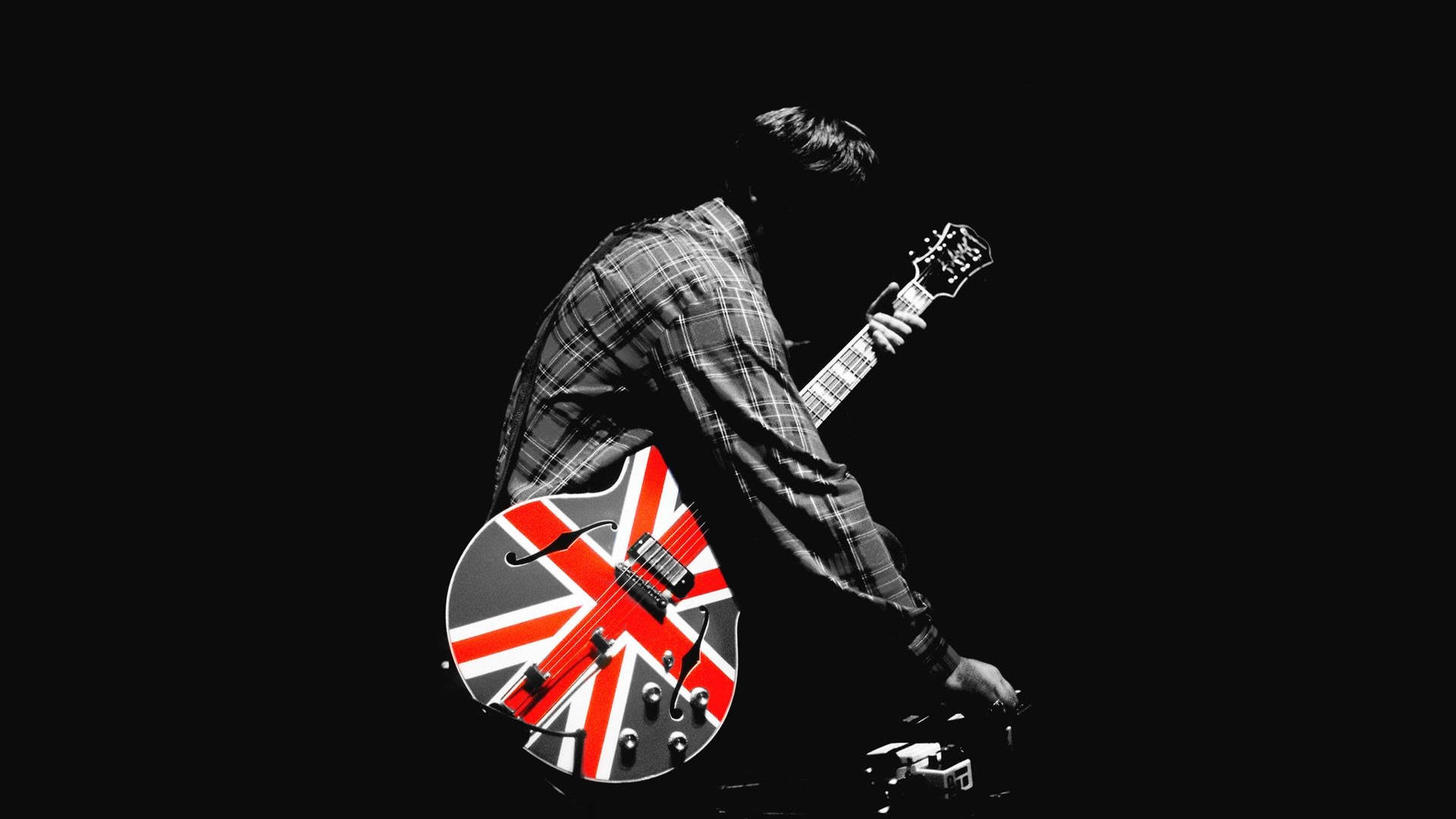 Oasis 吉他 壁紙 — 一位穿著格仔恤衫的音樂家，在純黑色背景下彈奏一把帶有醒目英國國旗圖案的結他。