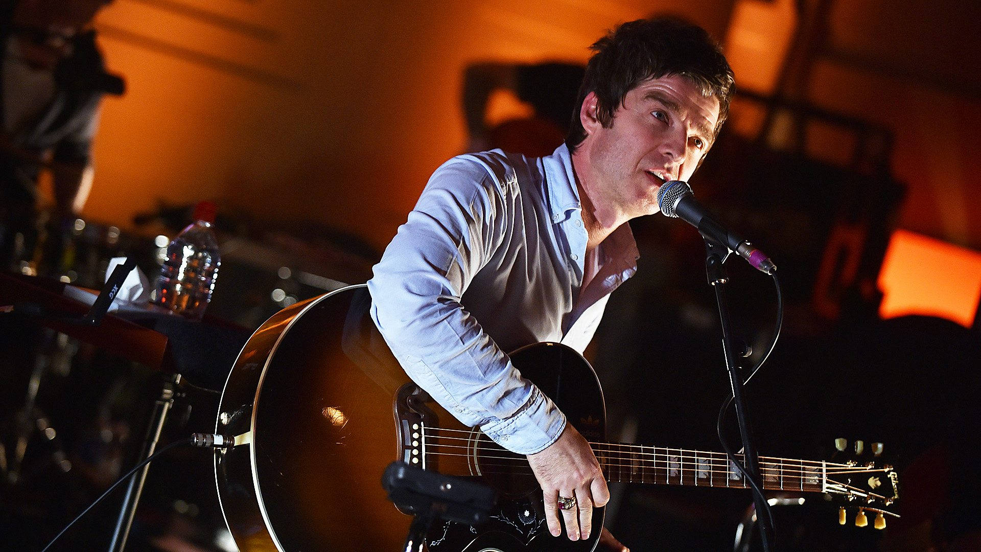 Oasis 吉他手 Noel Gallagher 壁紙 — 一位男音樂家在溫暖的舞台燈光下，一邊彈奏木結他，一邊熱情地對著咪高峰唱歌。