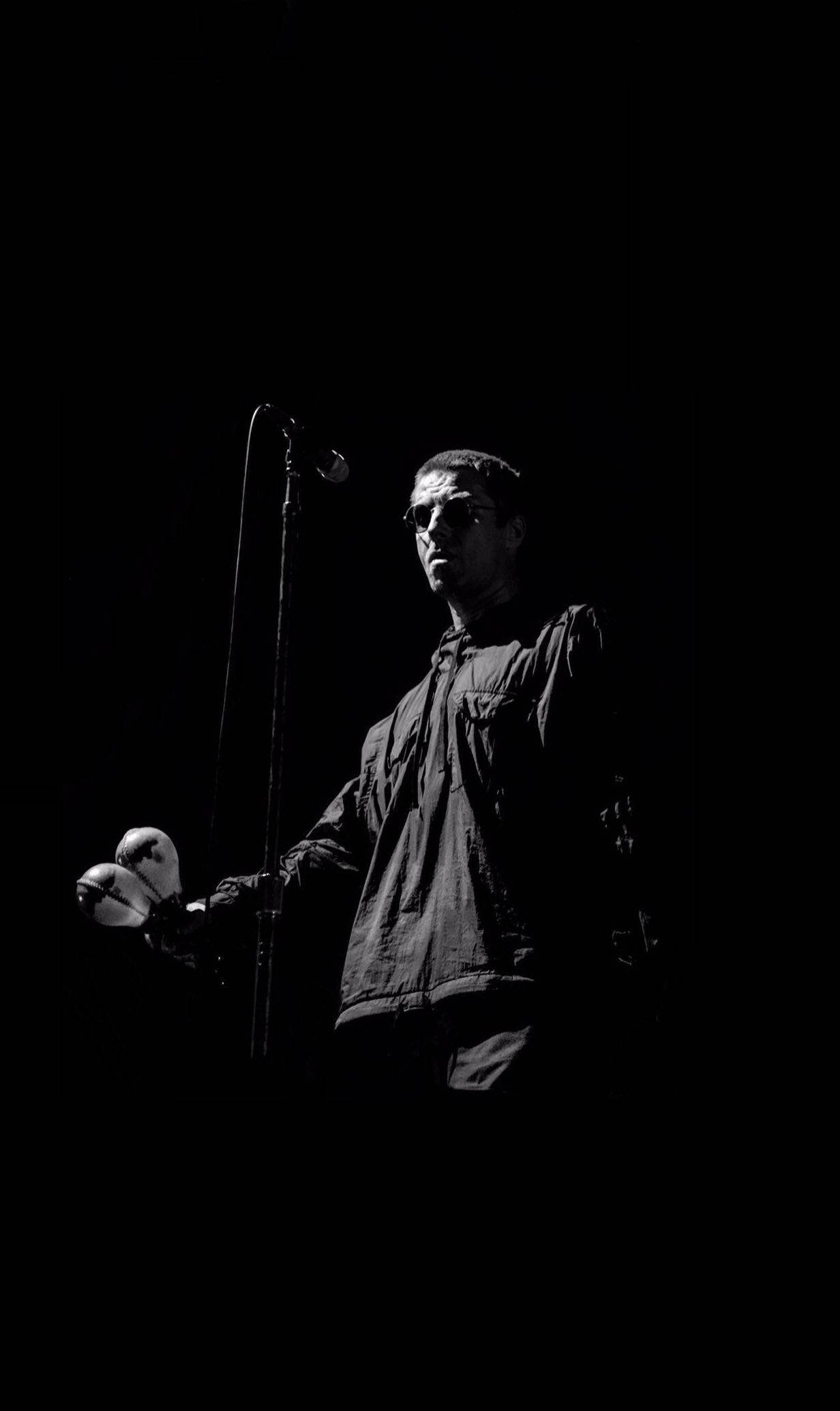 Oasis 持沙錘演唱 壁紙 — 一位戴著太陽眼鏡和外套的音樂家站在黑暗中，在戲劇性的聚光燈下拿著麥克風和沙槌。