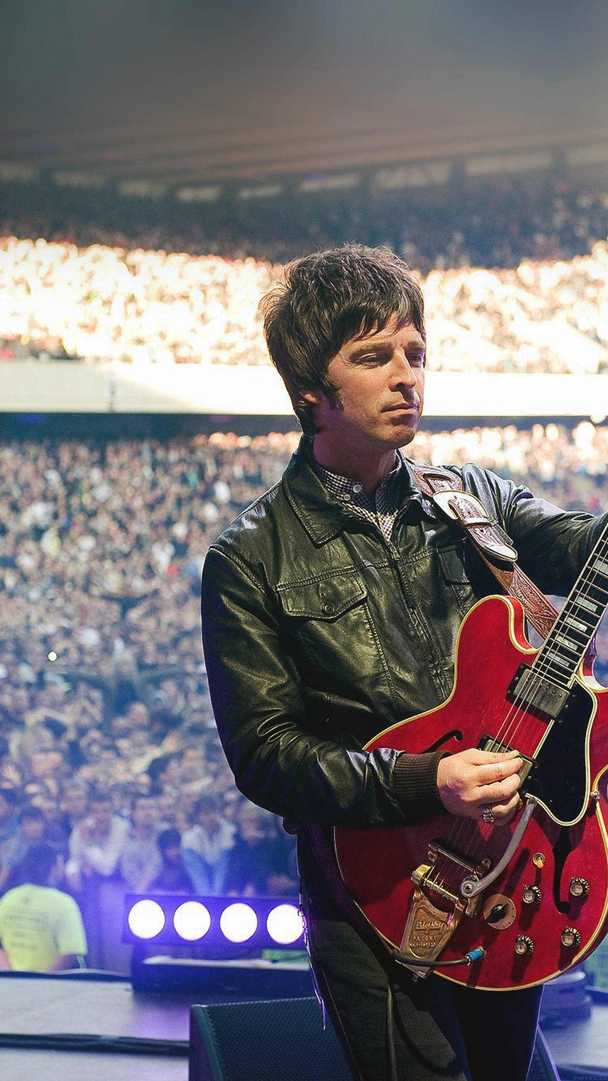 Oasis：諾爾彈吉他 壁紙 — 一位身穿黑色皮褸的音樂家在舞台上，在明亮的體育場燈光下，對著歡呼的龐大群眾彈奏紅色電結他。