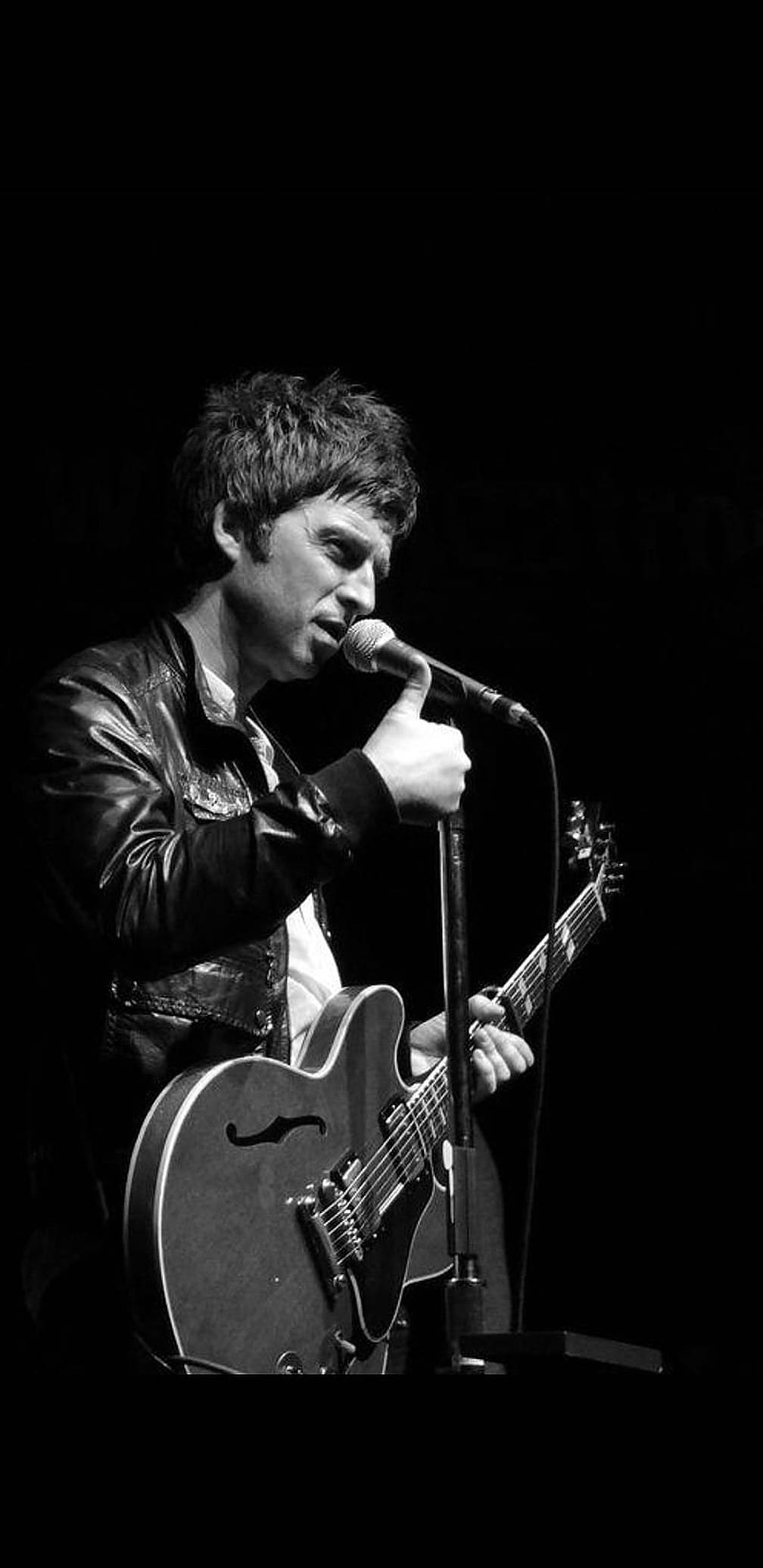 Oasis樂隊Noel Gallagher 壁紙 — 一位身穿皮褸的音樂家，在純黑背景下，一邊拿着電結他，一邊熱情地對着咪高峰唱歌。