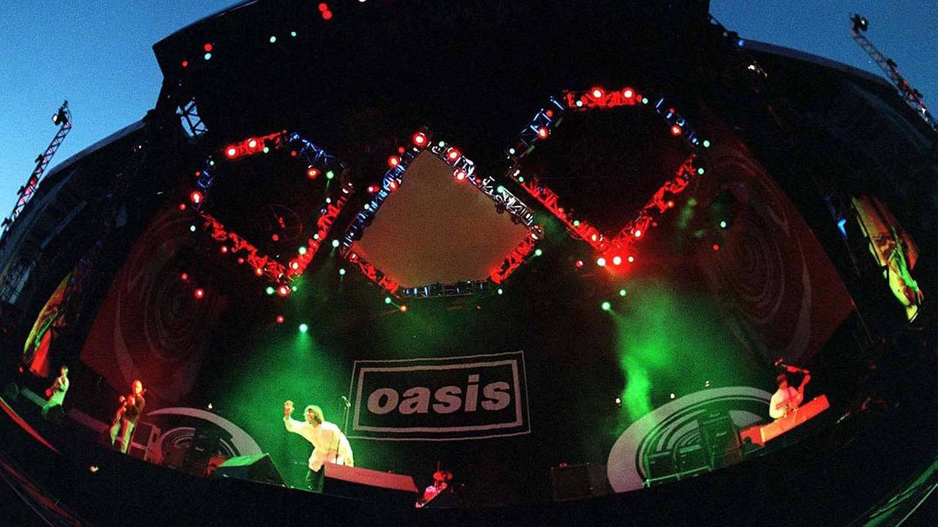 Oasis樂隊廣角舞台 壁紙 — Oasis 在充滿活力的舞台上表演的魚眼視角，舞台上有紅色和綠色的燈光，頭頂有一個心形燈架。