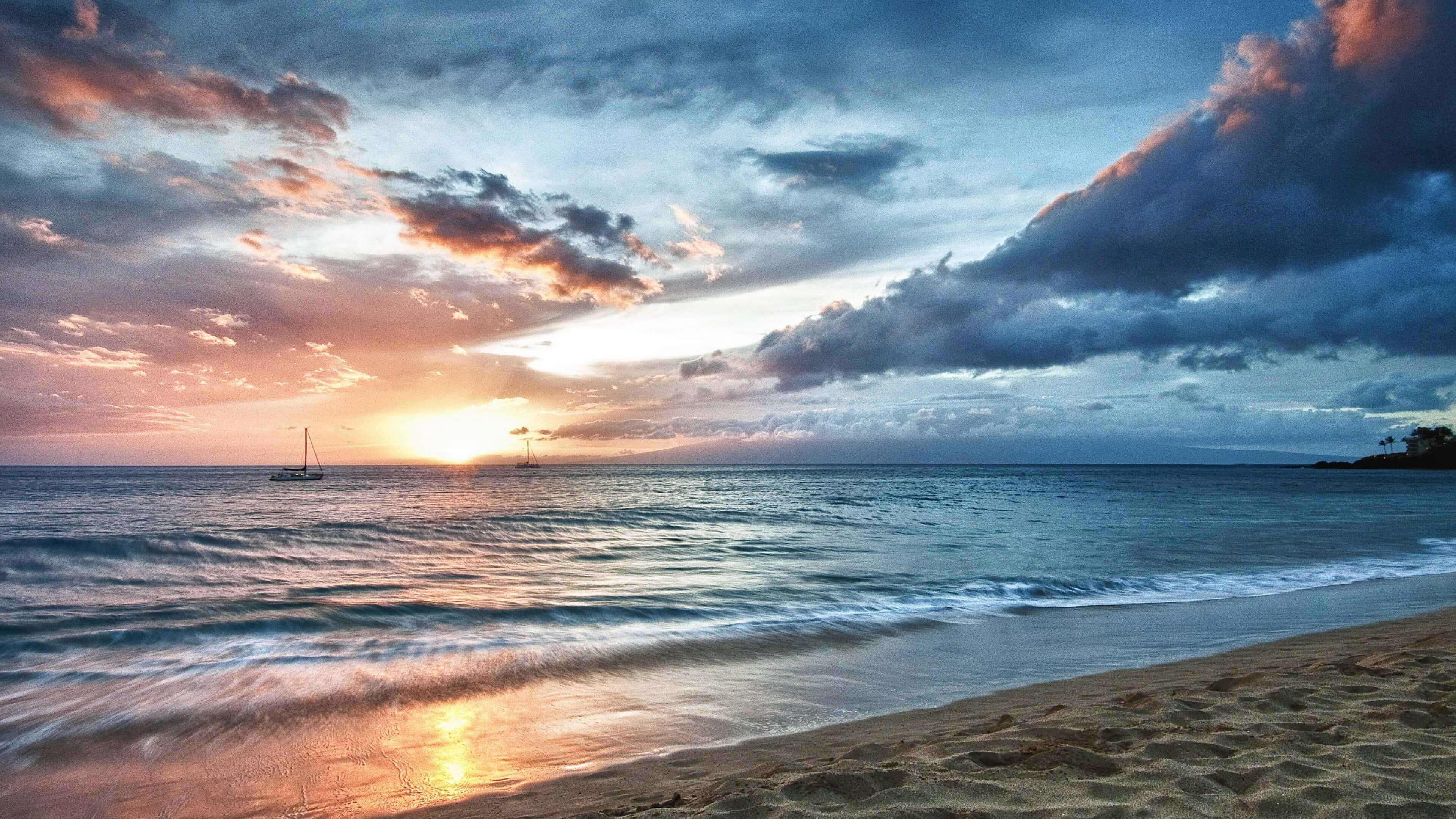 用 Ocean 4K iPad 接近大自然之美 壁紙 — 夕陽將天空染成火橙色和深藍色，帆船在沙灘附近平靜的海面上滑行。