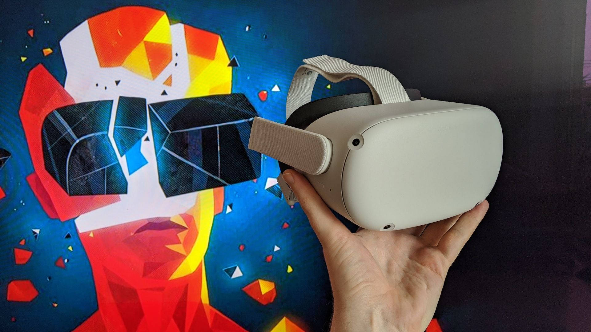 沉浸式遊戲體驗：Oculus Quest 2 壁紙 — 一隻手拿著白色VR頭戴式顯示器，對著一幅色彩鮮豔的幾何壁畫，壁畫上是一個風格化的臉孔，帶有鮮明的紅色、黃色和藍色形狀。