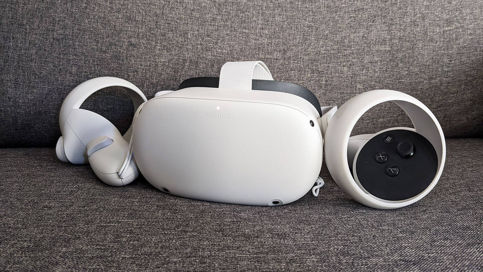 尊貴Oculus Quest 2 壁紙 — 一個白色Oculus VR頭戴式裝置和控制器整齊地放在一張有紋理的灰色布藝梳化上。