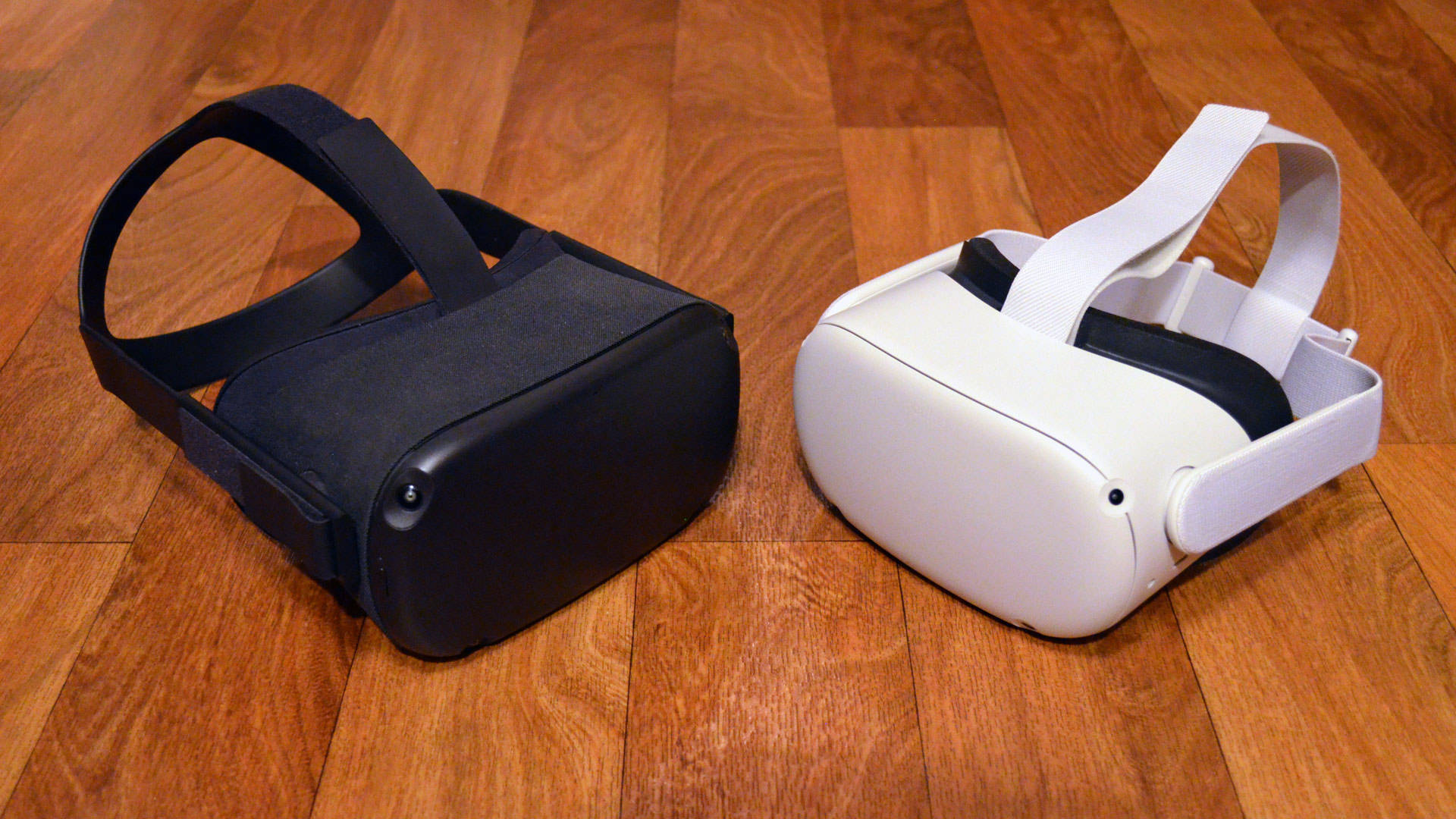 Oculus Quest 2 VR頭戴裝置 壁紙 — 兩個VR頭戴式顯示器，一黑一白，並排放在溫暖的木地板上。