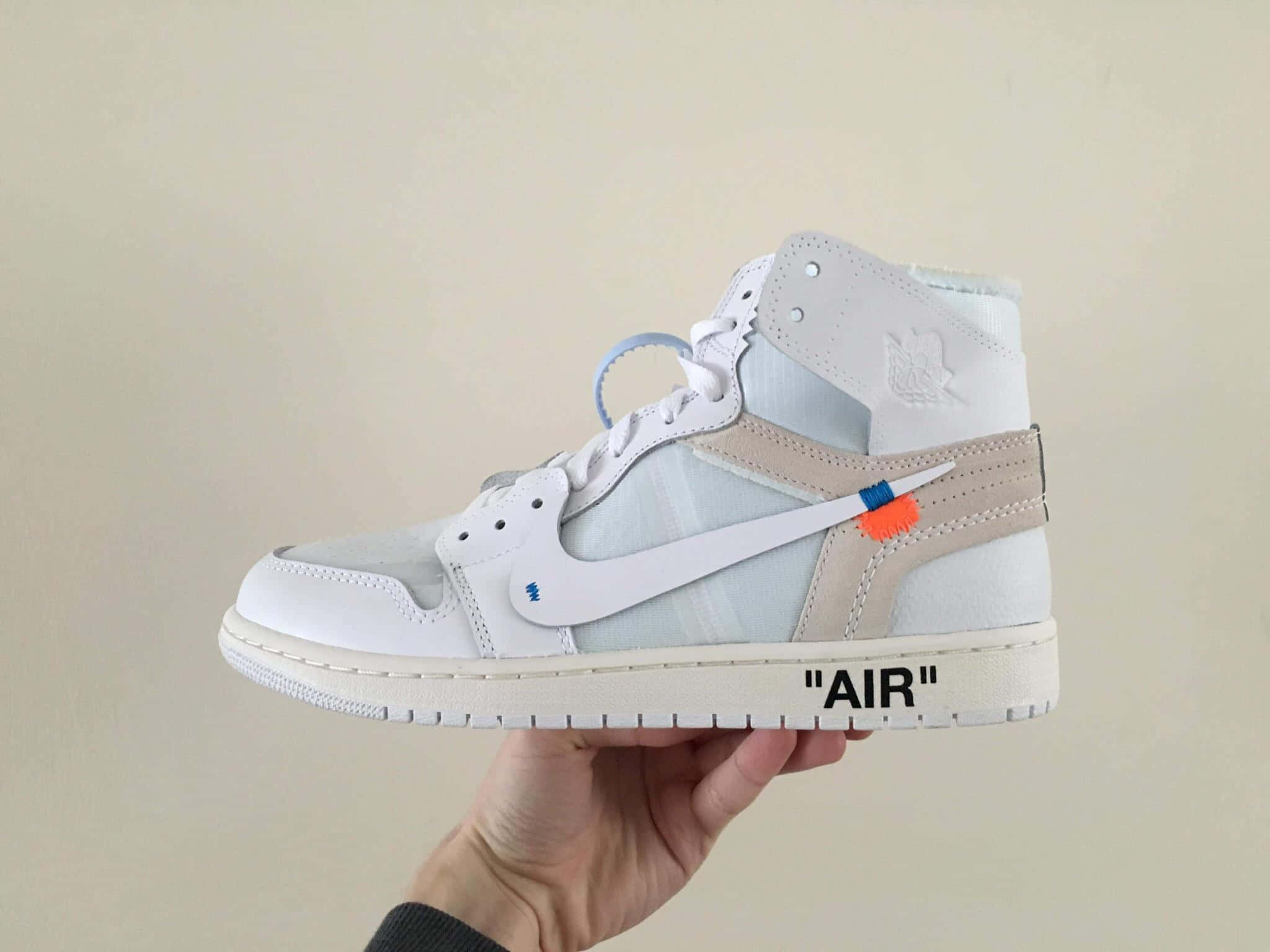 Off White 不可阻擋的時尚風格 壁紙 — 一隻手拿著一雙白色高筒運動鞋，帶有米色點綴和醒目的橙藍色品牌標誌，背景是純色米色牆壁。