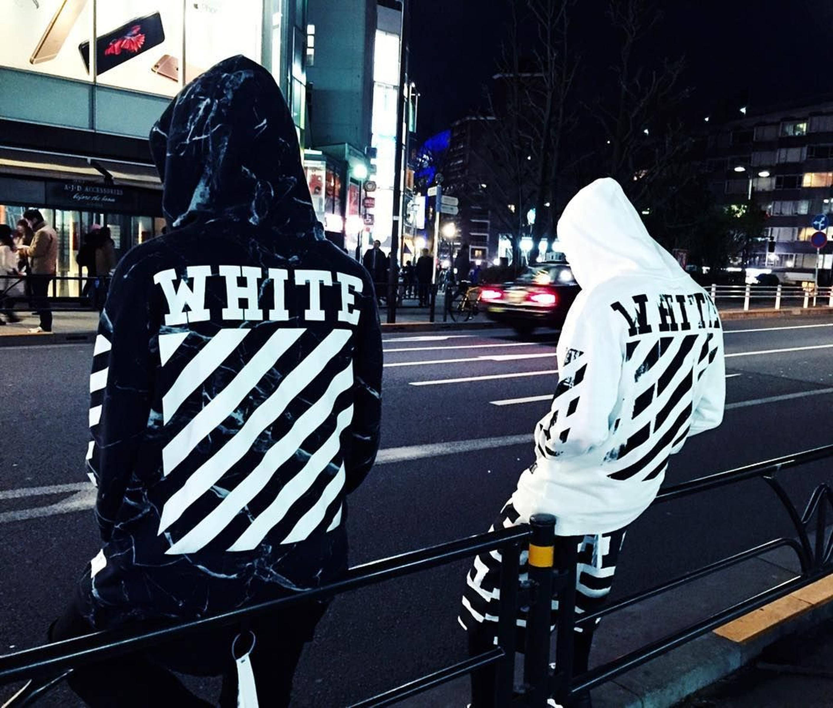 Off White 標誌夾克 壁紙 — 兩個穿著大膽黑白連帽衫的人坐在欄杆上，面向夜晚霓虹燈照亮嘅城市街道。