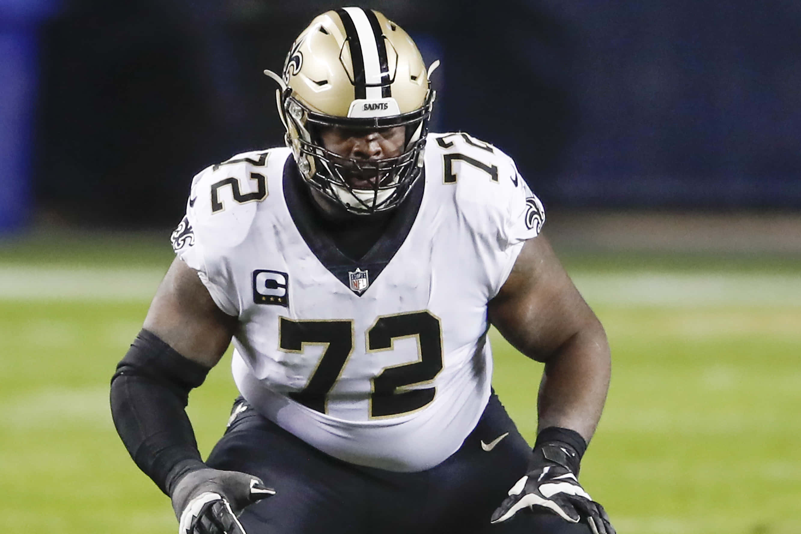 特龍·阿姆斯特德 美國職業橄欖球 Offensive Tackle 壁紙 — 新奧爾良聖徒隊的進攻鋒線球員身穿白色和金色球衣，在體育場燈光下於綠色足球場上激烈蹲伏。
