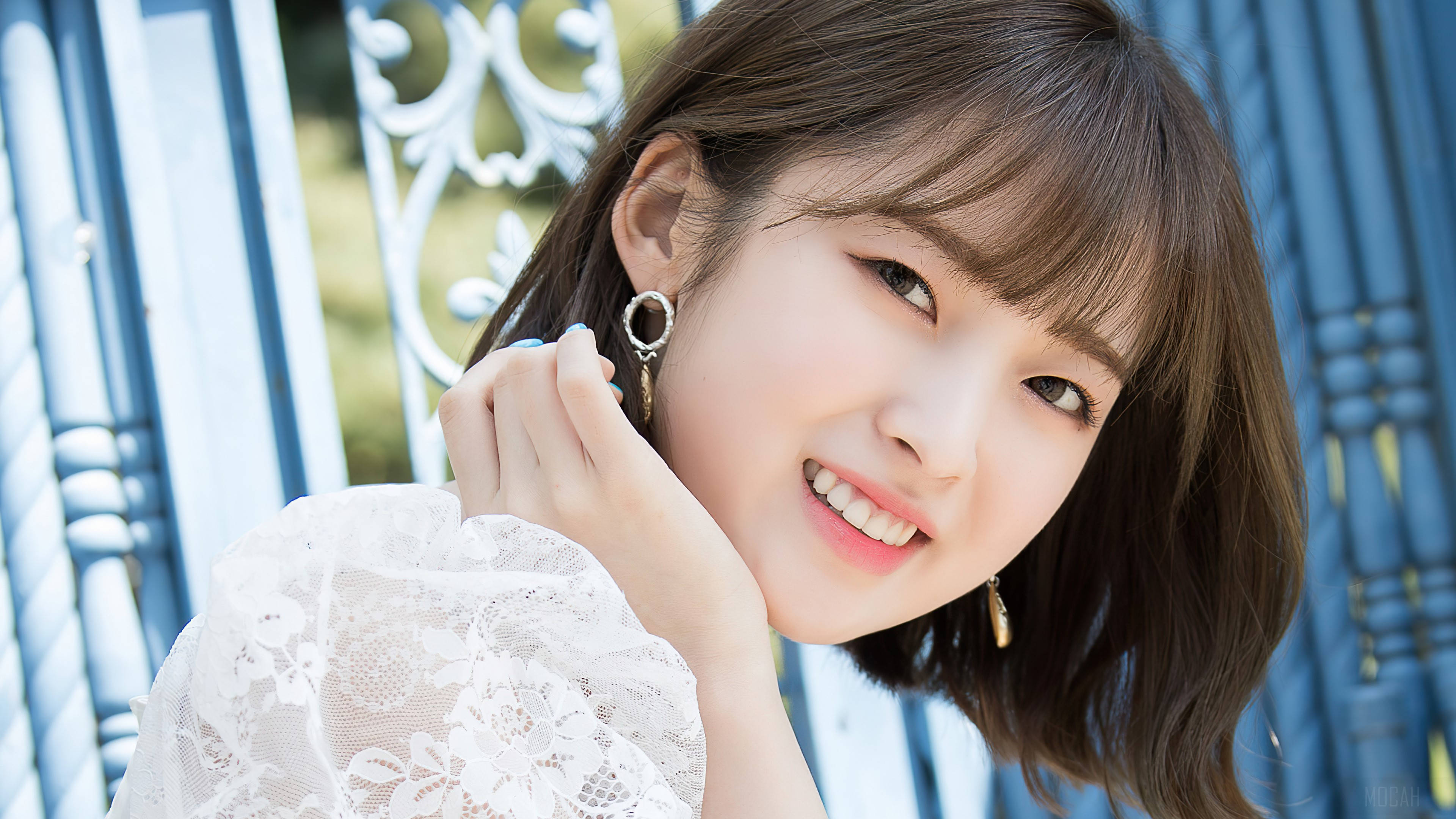 Oh My Girl Arin 純真笑容 壁紙 — 一位微笑的年輕女性，棕色頭髮，身穿白色蕾絲上衣，在晴朗的日子裡，在藍色裝飾金屬圍欄前擺姿勢。