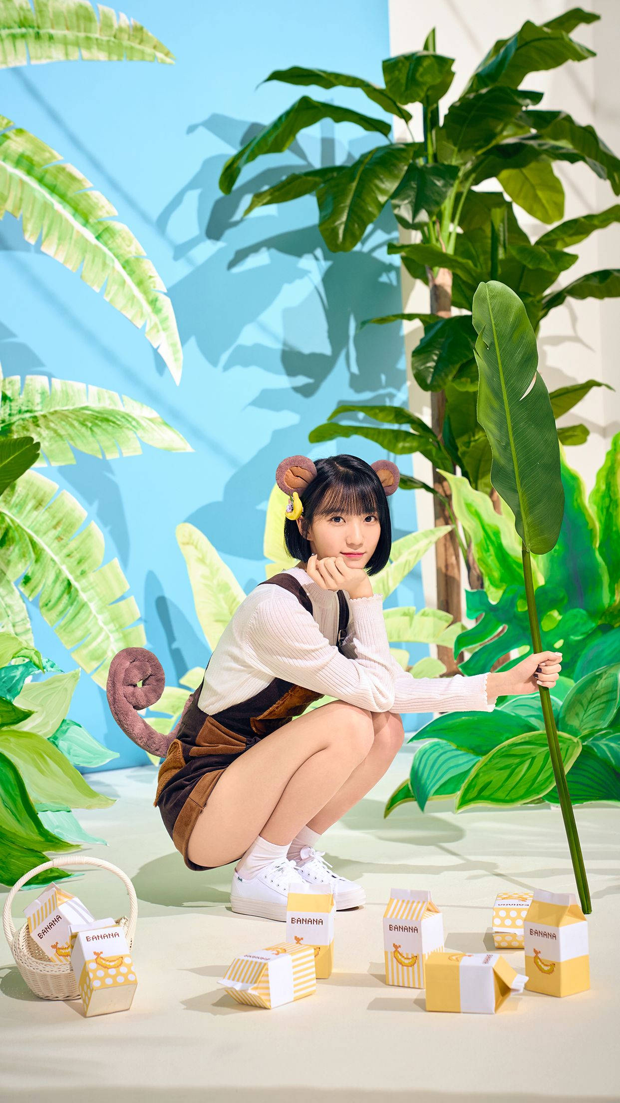 Oh My Girl Banhana Binnie 可愛造型 壁紙 — 一位戴著熊耳朵的年輕女性蹲在超大型熱帶葉子之間，旁邊是香蕉主題的盒子，背景是明亮的藍色牆壁。