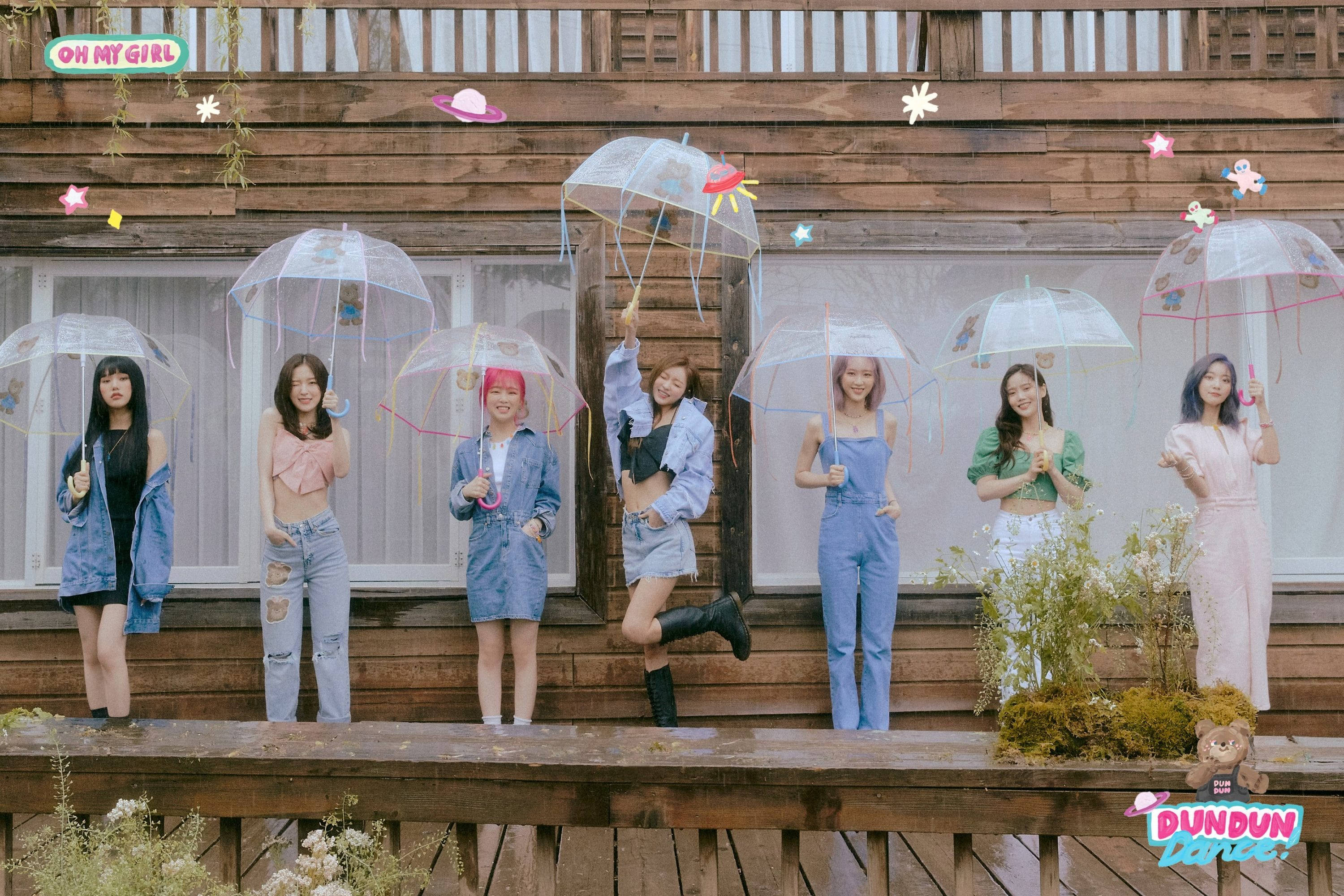 Oh My Girl《Dun Dun Dance》熱舞演出 壁紙 — 七個穿著色彩繽紛服裝的女孩在木甲板上愉快地擺姿勢，手持裝飾有俏皮裝飾的透明雨傘。