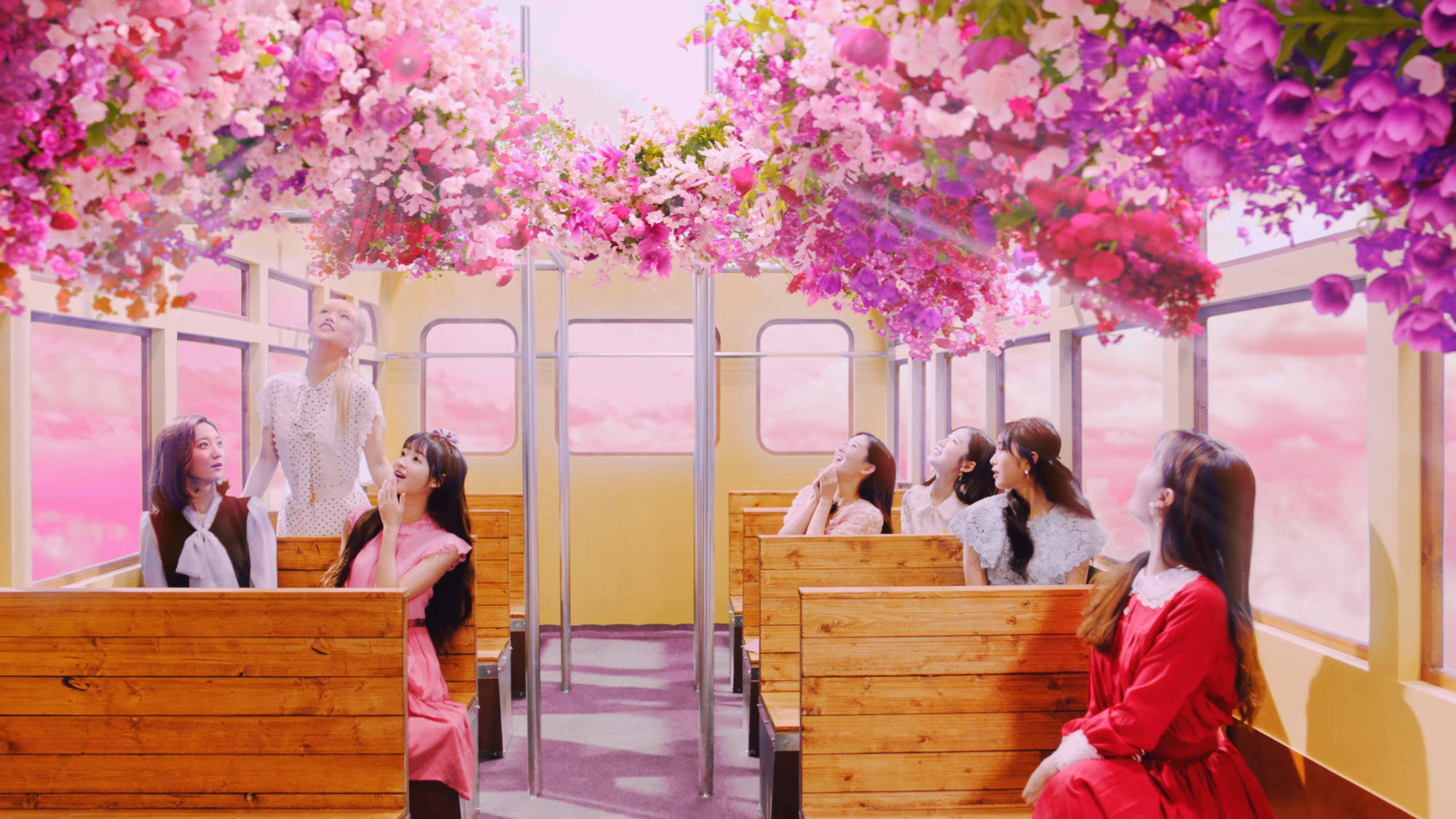 Oh My Girl 第五季音樂錄影帶夢幻列車 壁紙 — 年輕女性在復古火車車廂內向上仰望，鮮豔的粉紅色和紫色花朵從天花板如瀑布般落下，映襯著粉彩色的天空。