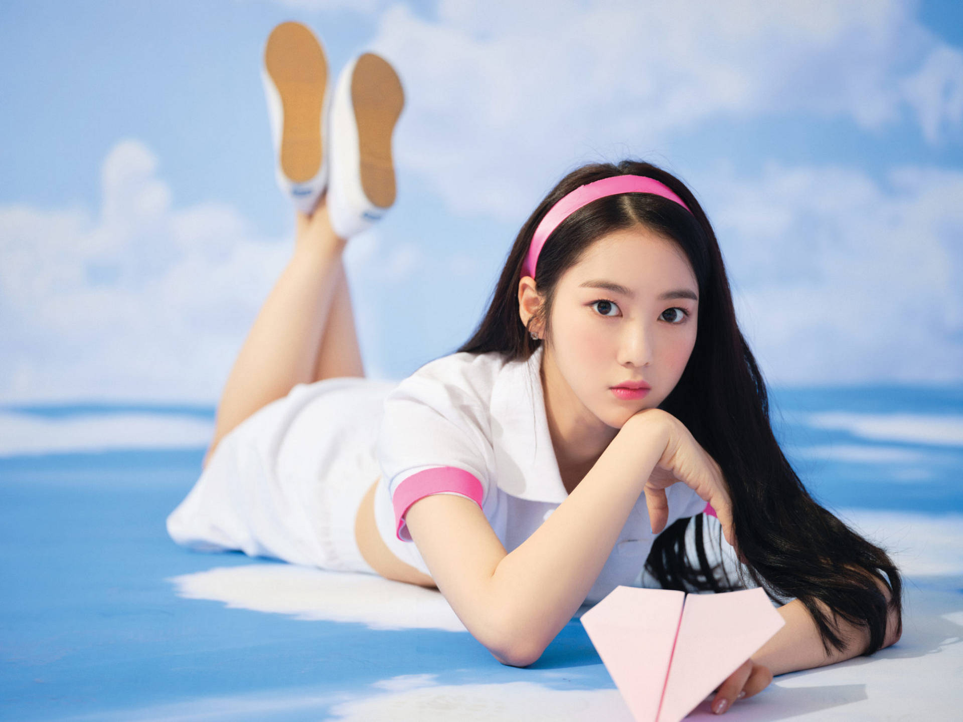 Oh My Girl Jiho 紙飛機造型 壁紙 — 一位身穿白色服裝帶有粉紅色點綴的年輕女子躺在藍色表面上，手持粉紅色紙飛機，背景是陰天天空。
