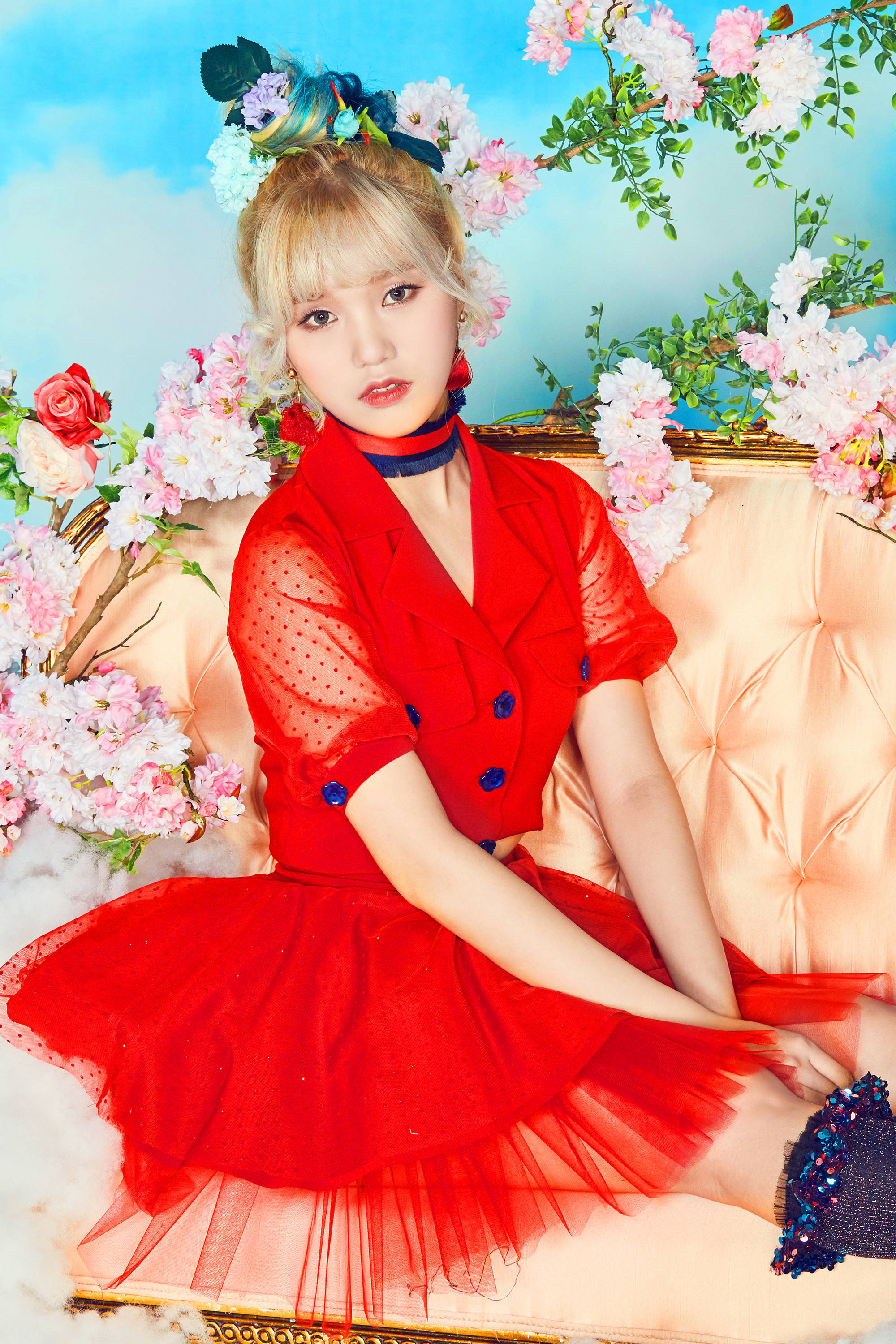 Oh My Girl Mimi 穿紅色服裝 壁紙 — 一位身穿鮮艷紅裙的年輕女士，優雅地坐在一張簇絨蜜桃色梳化上，周圍是盛開的粉紅和白色花朵，背景是柔和藍天。