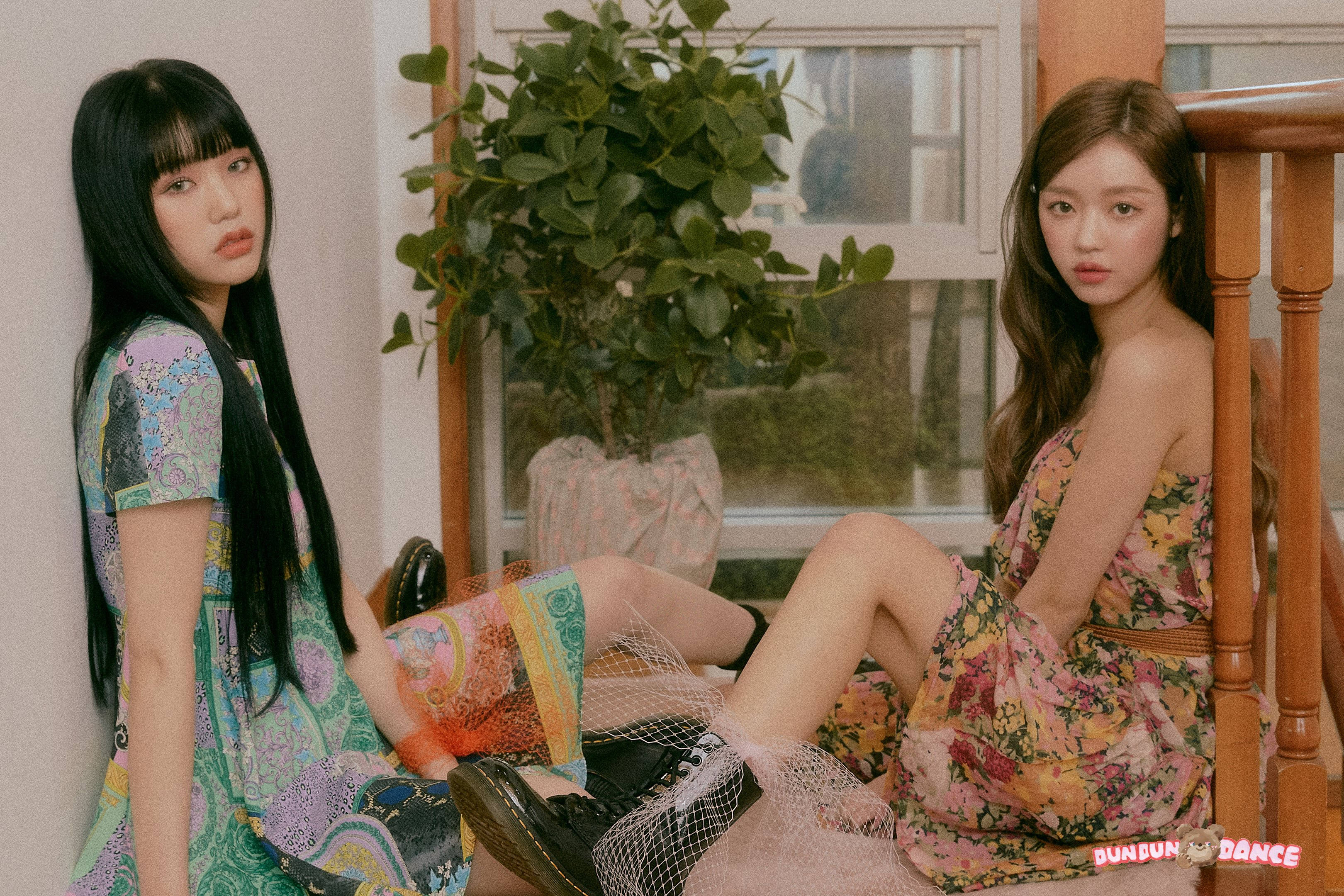 Oh My Girl Mimi與YooA 花朵對坐 壁紙 — 兩位穿著花卉連衣裙的年輕女性坐在室內靠近窗邊，窗邊有一盆植物，她們在柔和自然光線下看著相機。