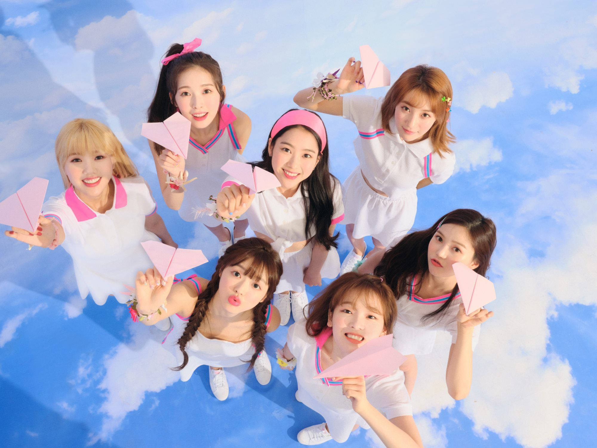 Oh My Girl 拿紙飛機的可愛畫面 壁紙 — 七位微笑的年輕女性身穿白色服裝，在藍天白雲下手持粉紅色紙飛機。