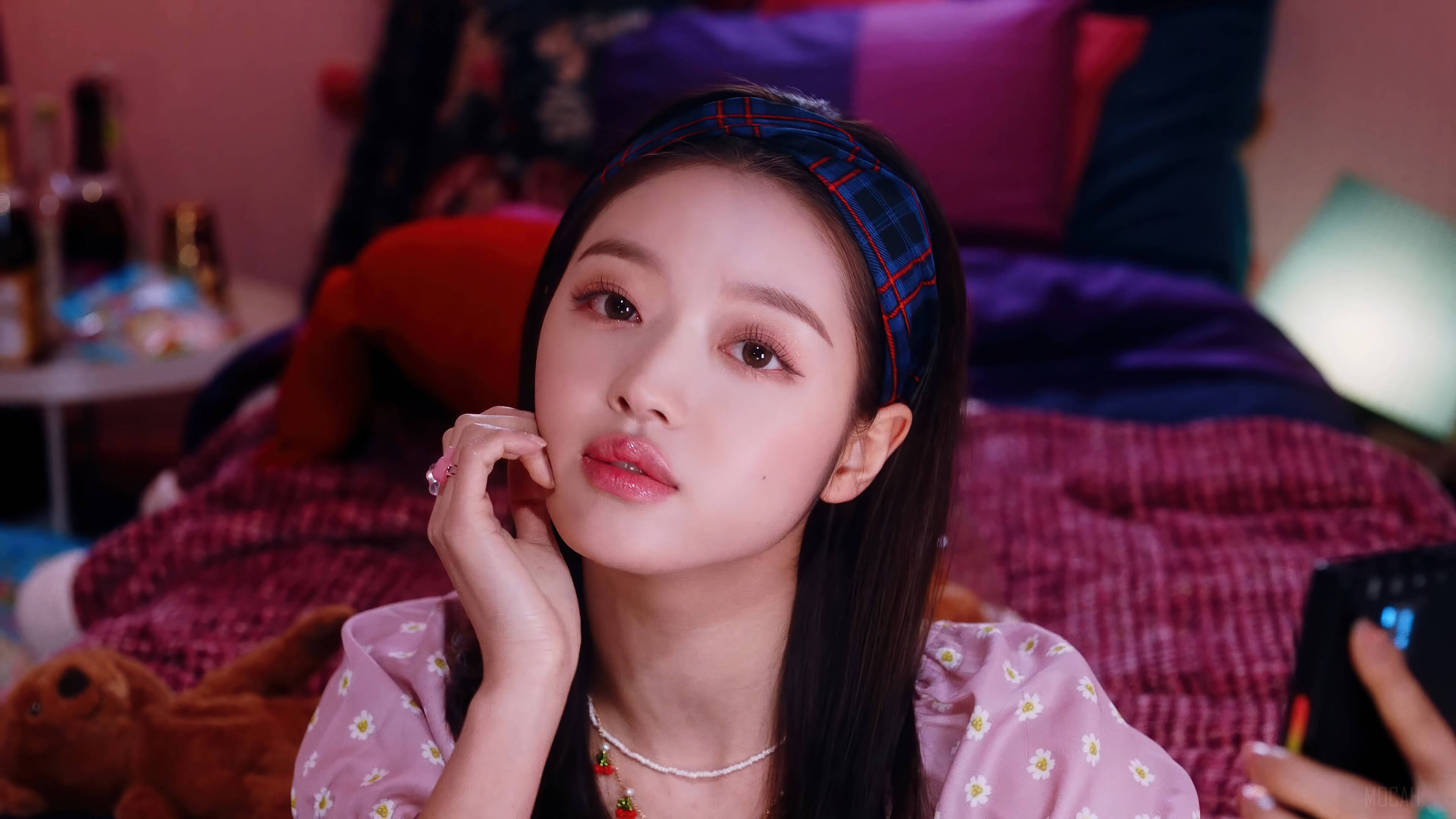 OH MY GIRL YooA 粉紅造型 壁紙 — 一位戴著格仔頭帶的年輕女子，將下巴擱在手上，躺在一張有彩色枕頭和泰迪熊的床上。
