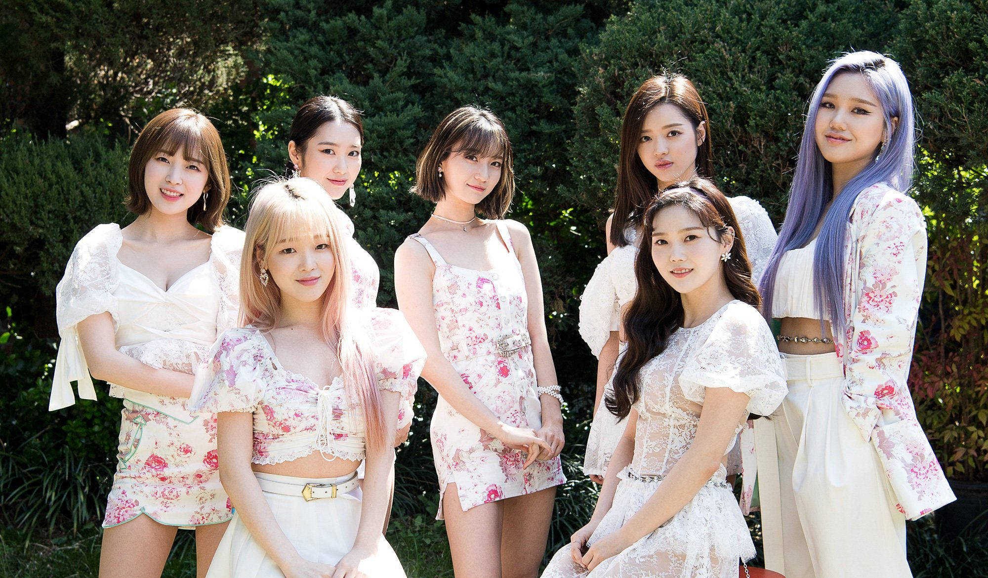 OH MY GIRL 白色花卉蕾絲造型 壁紙 — 七位年輕女性身穿花卉白色服裝，在陽光明媚的花園中一起擺姿勢，身後是茂密的綠色灌木叢。