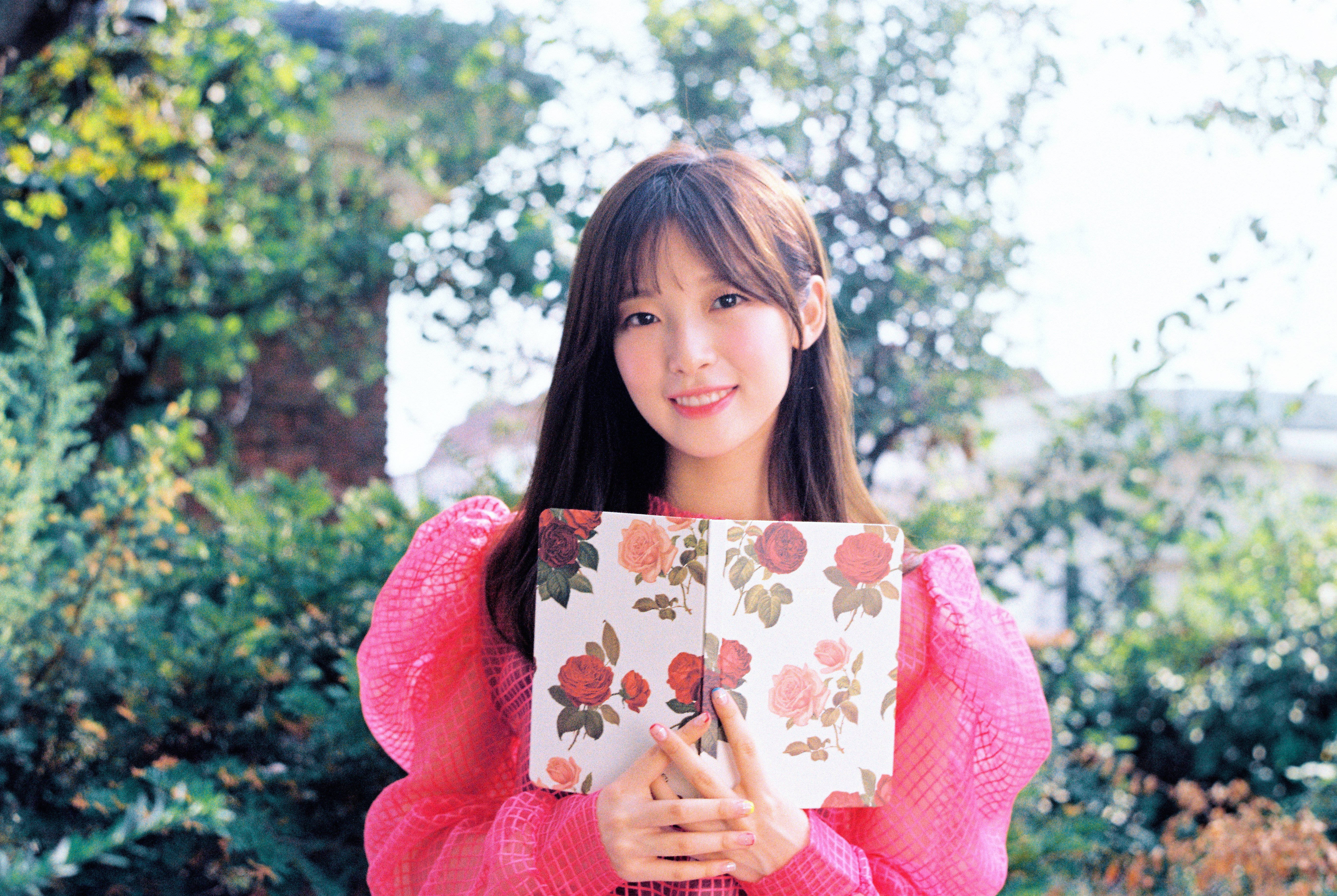 Oh My Girl 美麗的Arin 壁紙 — 一位微笑的年輕女性身穿亮粉紅色網眼上衣，在戶外茂密的綠植中手持花卉圖案筆記本。