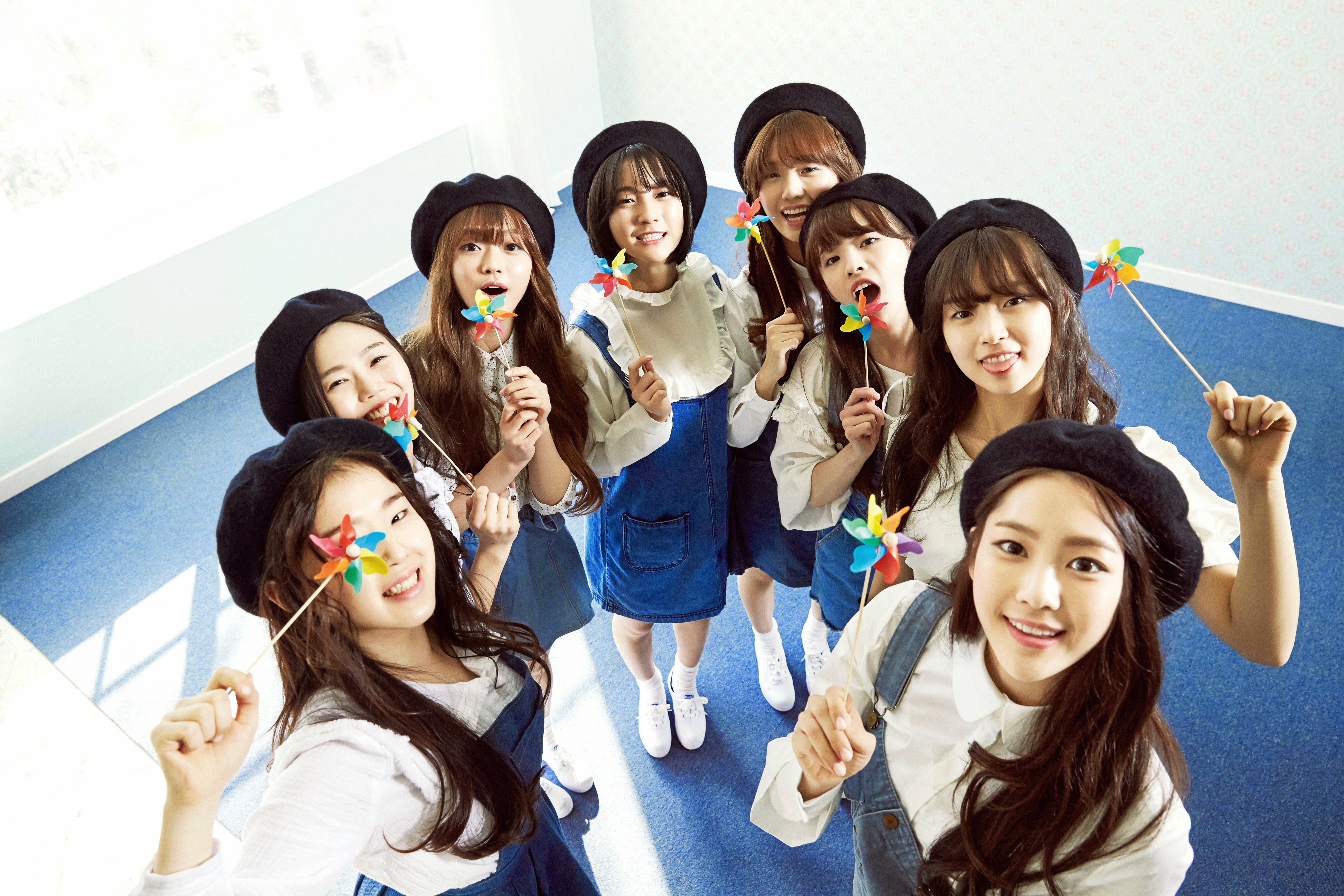 Oh My Girl 藍色連身裙造型 壁紙 — 七位微笑的年輕女性戴著黑色貝雷帽和牛仔工裝褲，在明亮的室內走廊裡愉快地拿著彩色風車擺姿勢。
