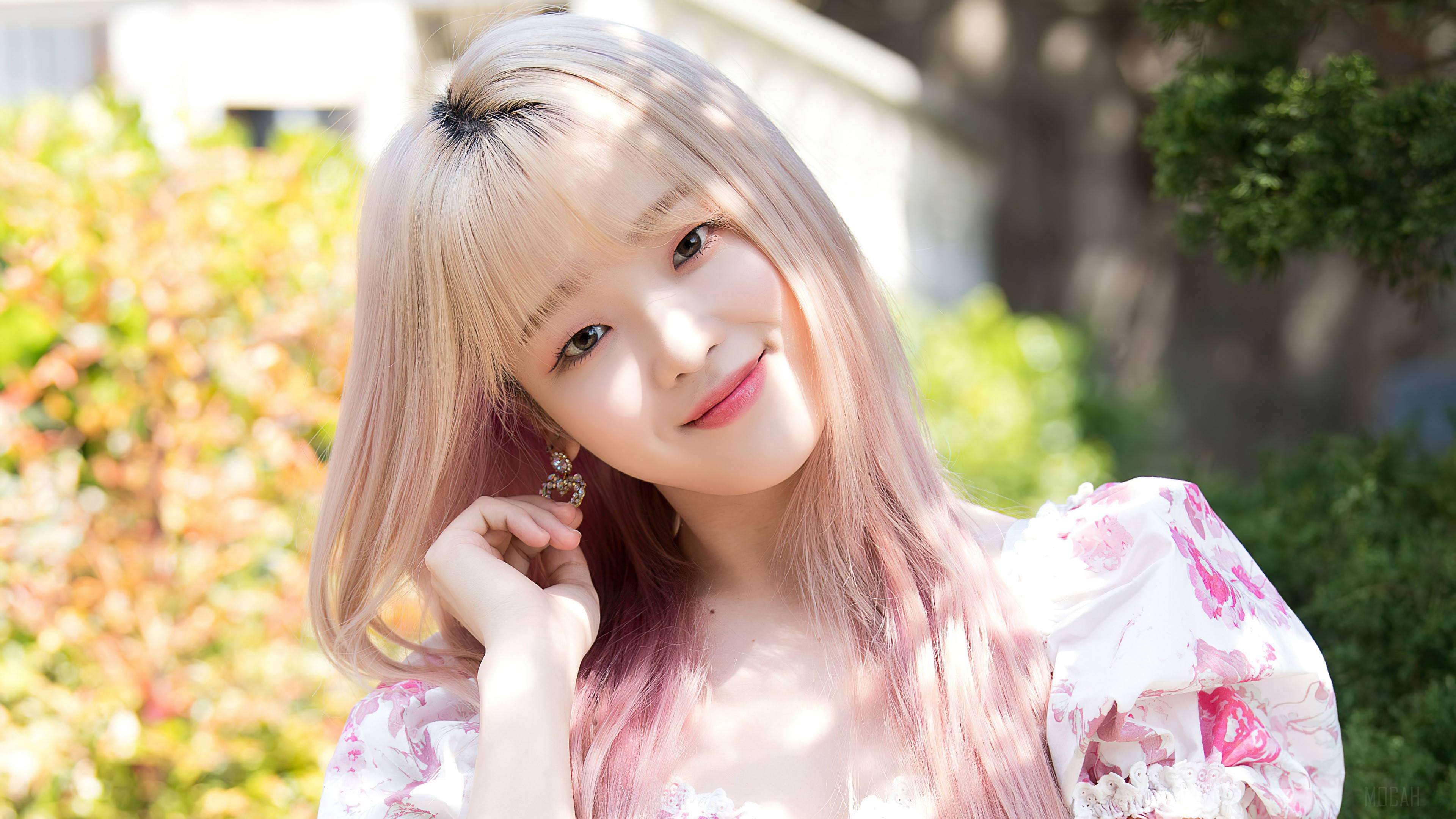 Oh My Girl 魅力Seunghee 壁紙 — 一位粉紅色頭髮、身穿花卉連衣裙的年輕女性在陽光普照的花園樹葉中輕柔微笑。