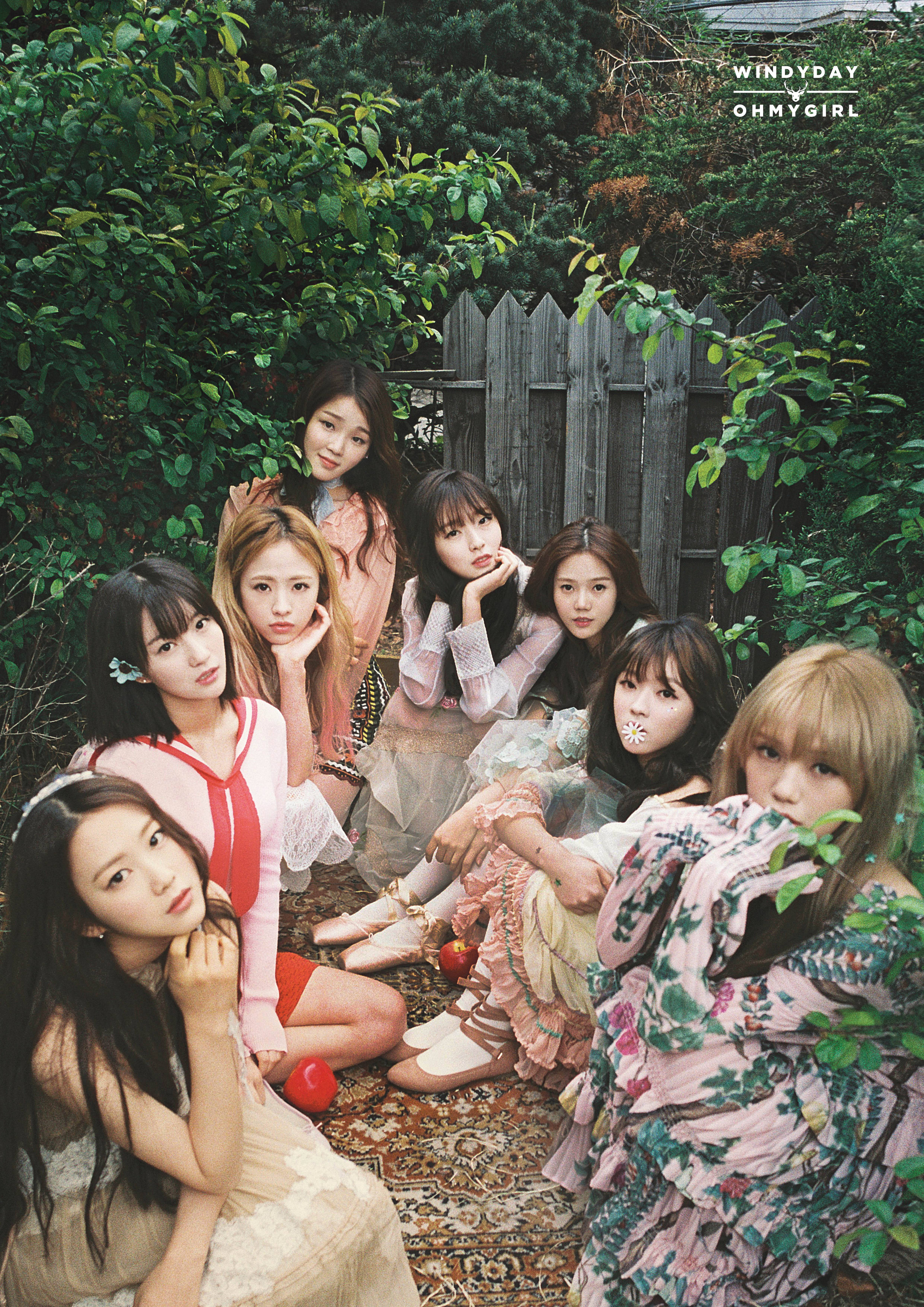 Oh My Girl《Windy Day》MV畫面 壁紙 — 七位年輕女性身穿柔和花卉服裝，在圖案地毯上若有所思地擺姿勢，周圍是茂密的綠色植物和風化的木柵欄。