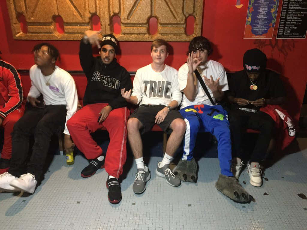 Ohgeesy 與 Shoreline Mafia 坐紅沙發 壁紙 — 五位年輕男性穿著休閒街頭服飾，在光線昏暗的室內空間中，以有紋理的牆壁為背景，在紅色長椅上擺姿勢。