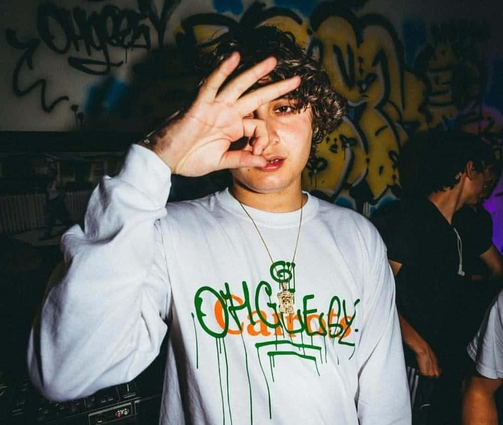 Ohgeesy - 西海岸嘻哈之王 壁紙 — 一個年輕嘅人，有曲髮，喺昏暗燈光下，喺一個佈滿塗鴉嘅牆前面做咗個眼神動作。