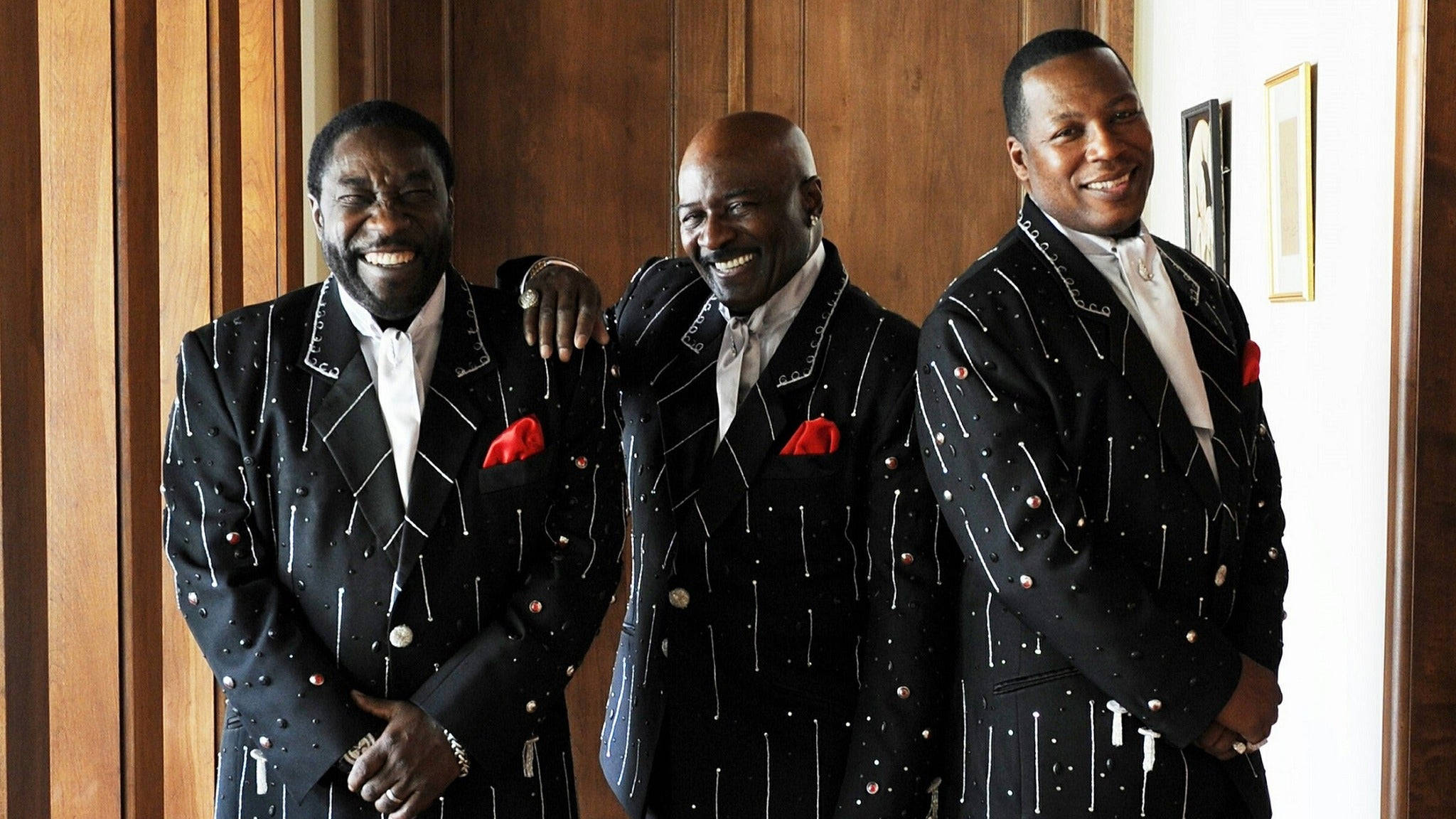 Ojays 演唱會門票 Hollywood 壁紙 — 三位微笑的男士穿著相配的黑色亮片西裝，搭配紅色口袋方巾，在木鑲板牆前溫馨地擺姿勢。