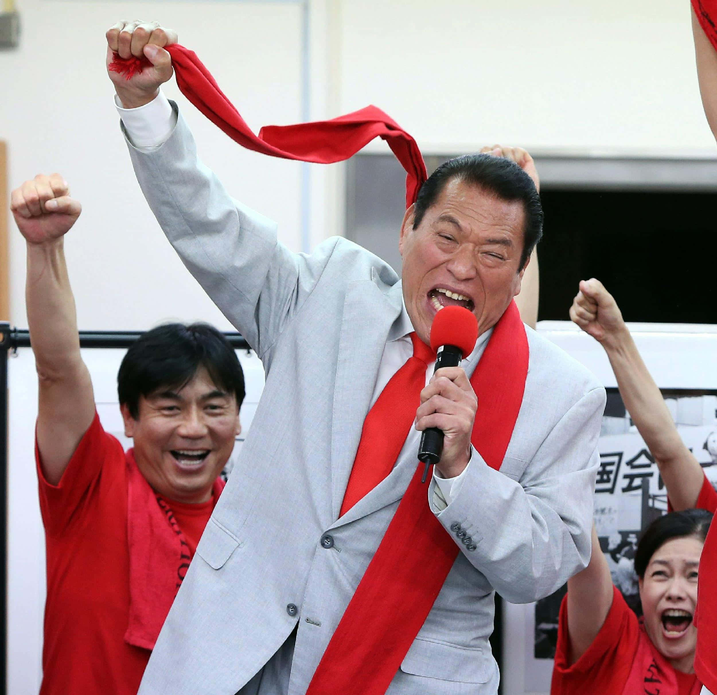 安東尼奧·因oki主導職業摔角賽事 壁紙 — 一名身穿灰色西裝、打紅色領帶的男子對著麥克風大喊，同時高舉紅絲帶和拳頭，在歡呼的支持者中慶祝勝利。