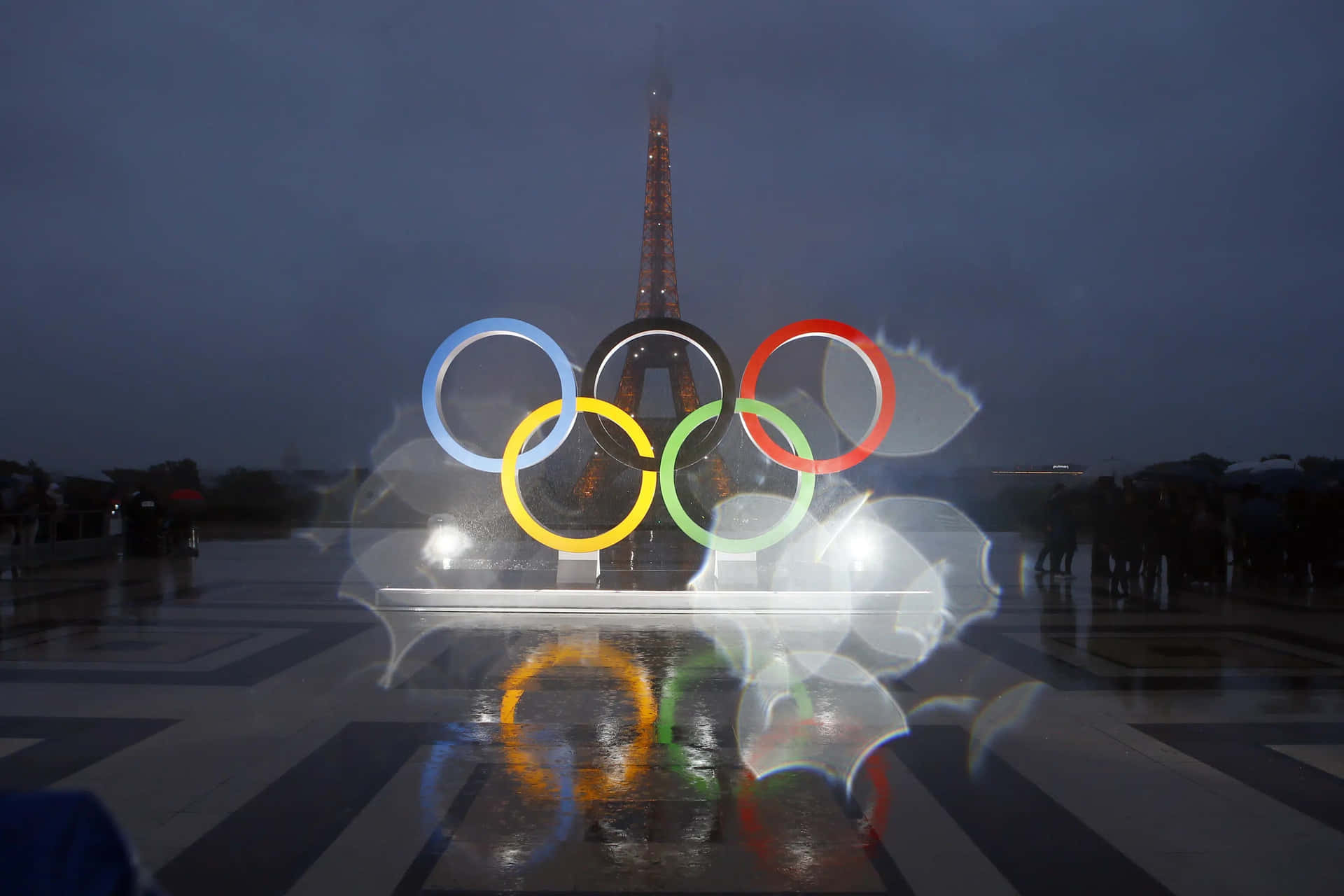 夜間奧運五環亮起，背景為艾菲爾鐵塔，濕漉漉的人行道反射光線。