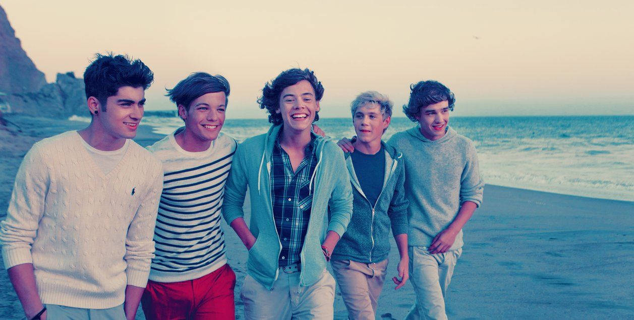 One Direction 海灘熱舞 壁紙 — 五位微笑的年輕男子在沙灘上一起擺姿勢，身後是海浪和懸崖，天空呈現柔和的暮色。