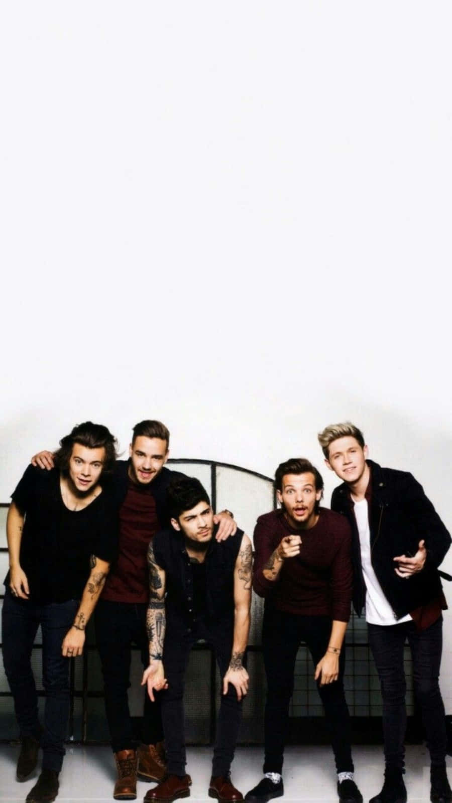 One Direction 白底男孩團 iPhone 壁紙 — 五名身穿深色休閒服的年輕男子在一個純白色牆壁前擺出俏皮的姿勢，身後有金屬欄杆。
