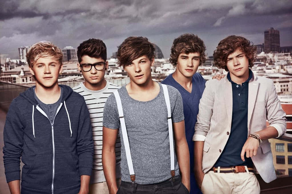 以 One Direction 筆記本電腦展開數位冒險之旅 壁紙 — 五位年輕男子穿著休閒服裝，在陰雲密布的城市天際線背景下自信地擺姿勢。